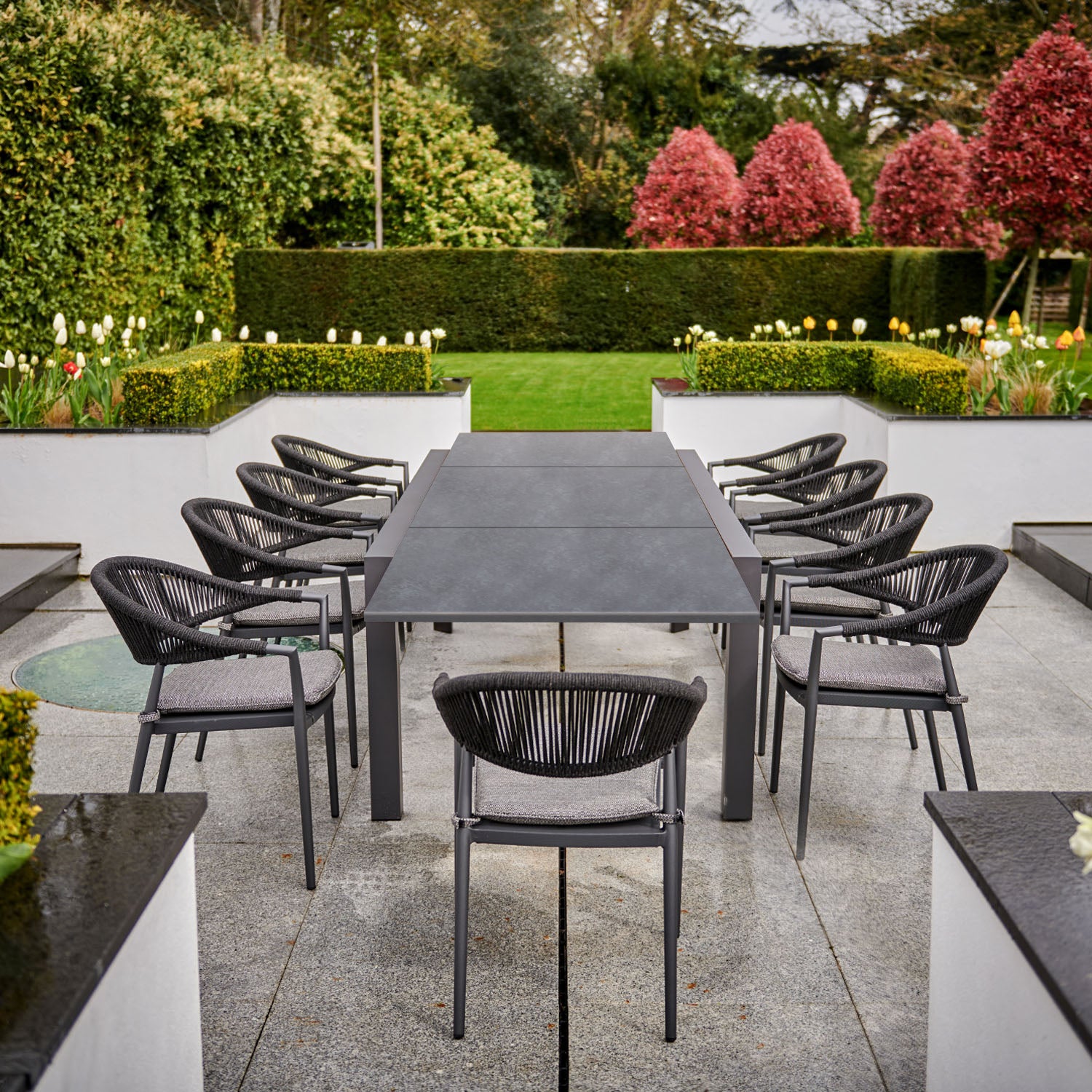 The Cloverly 8 Seat Rectangular Extending Dining Set with Ceramic Table in Charcoal, crafted with weather-proof aluminum, features a rectangular table and eight black chairs adorned with removable sun-proof cushions, all surrounded by greenery and red blooming trees.