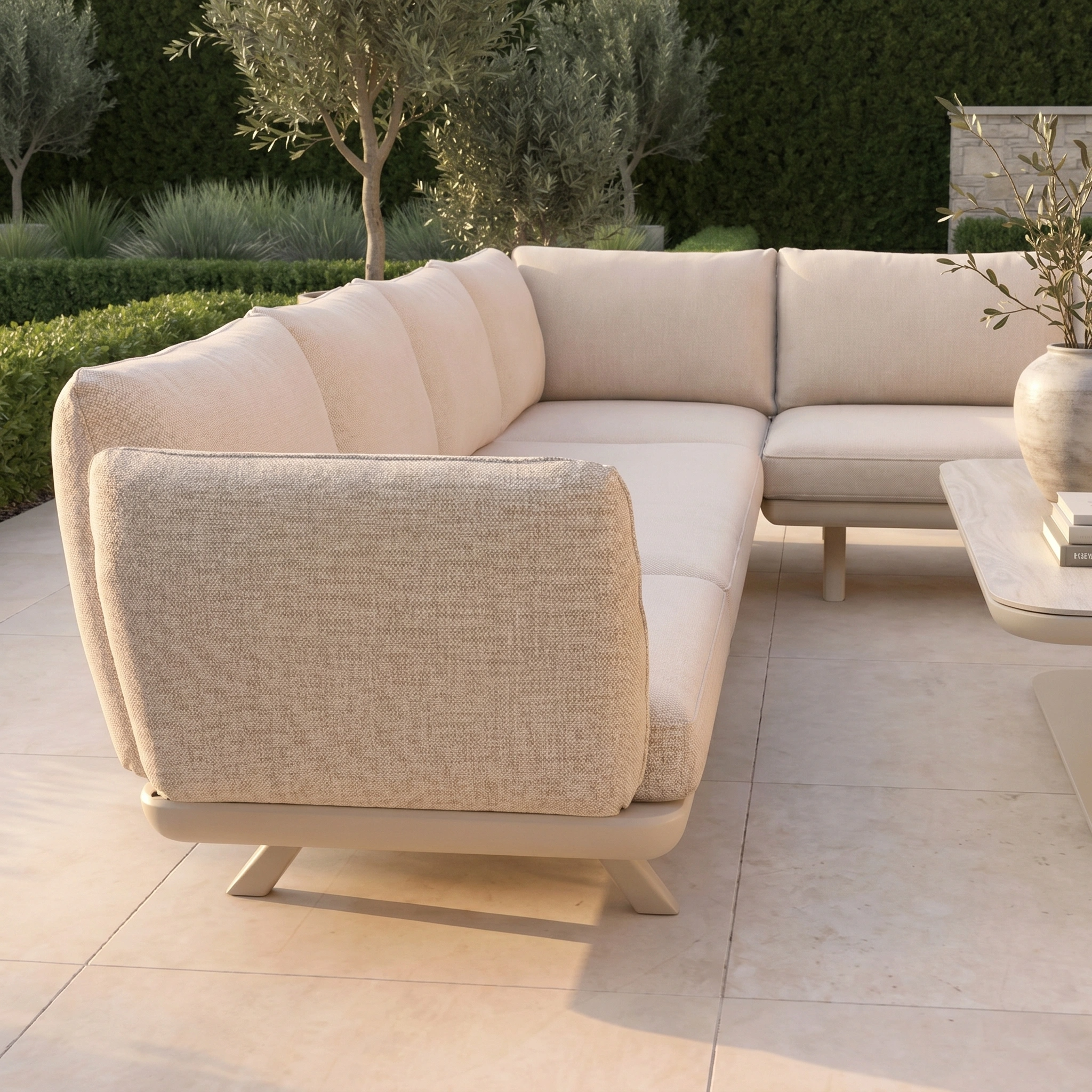 The Como U-Shape Corner Sofa Set with Altura Rising Table in Oatmeal sits on a stone patio, surrounded by greenery and small trees, featuring stylish aluminium outdoor furniture and a versatile rising table.