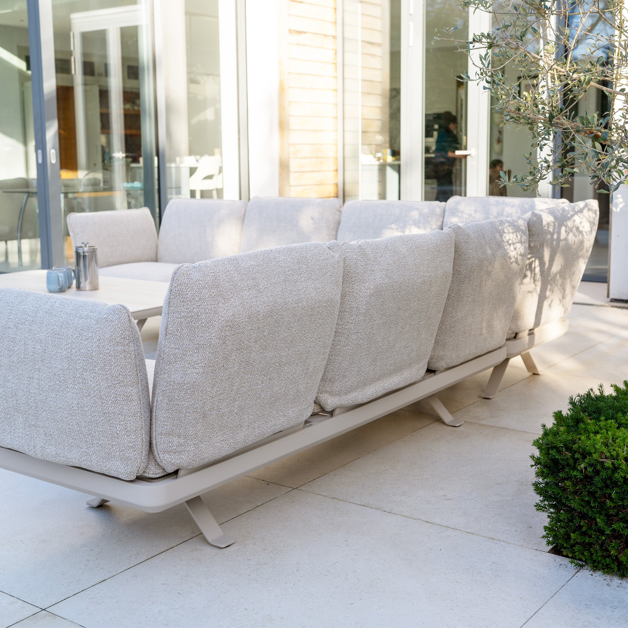 Como Corner Sofa Set with Altura Rising Table in Oatmeal, displayed on a patio surrounded by glass doors and lush greenery.