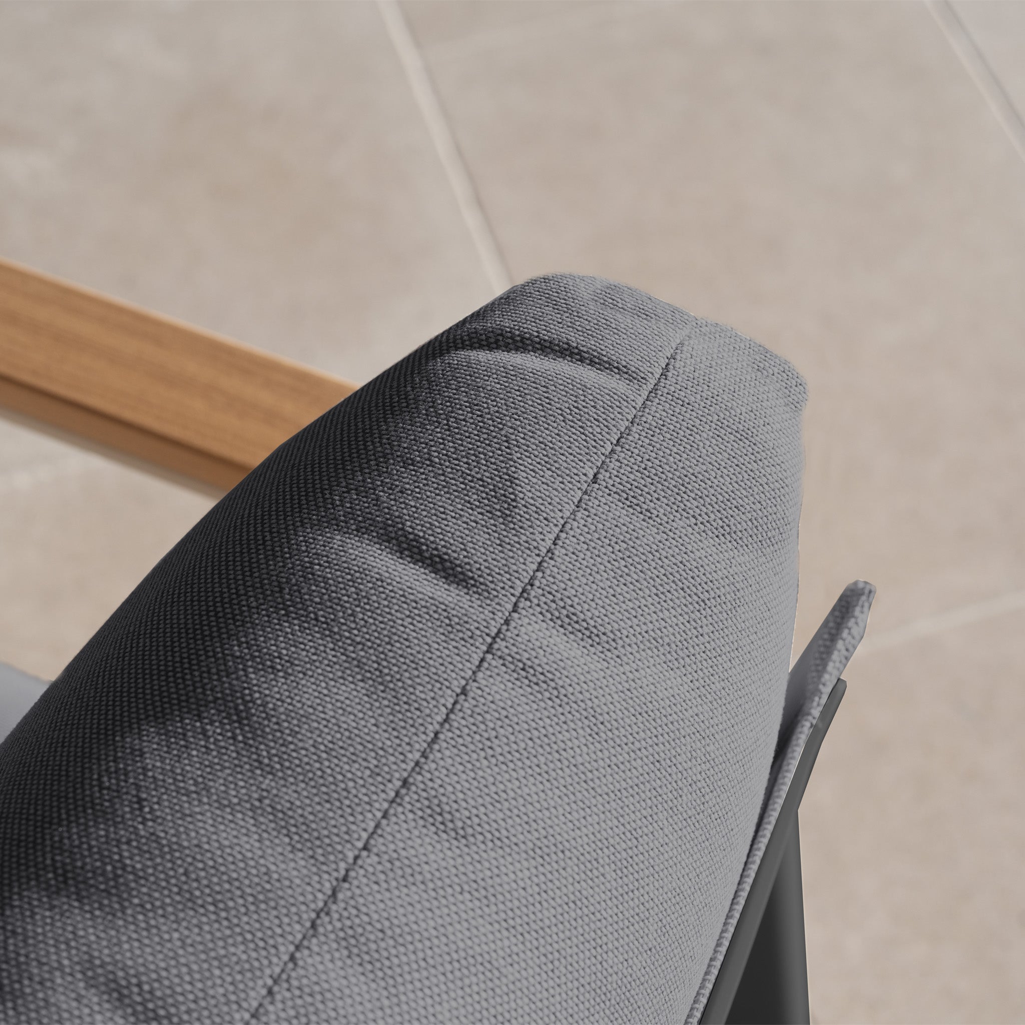 Close-up of a gray cushion on a chair with a wooden armrest and tiled floor, from the weather-resistant Cora 3 Seat Sofa Set in Grey—perfect for creating a modern outdoor sofa look.