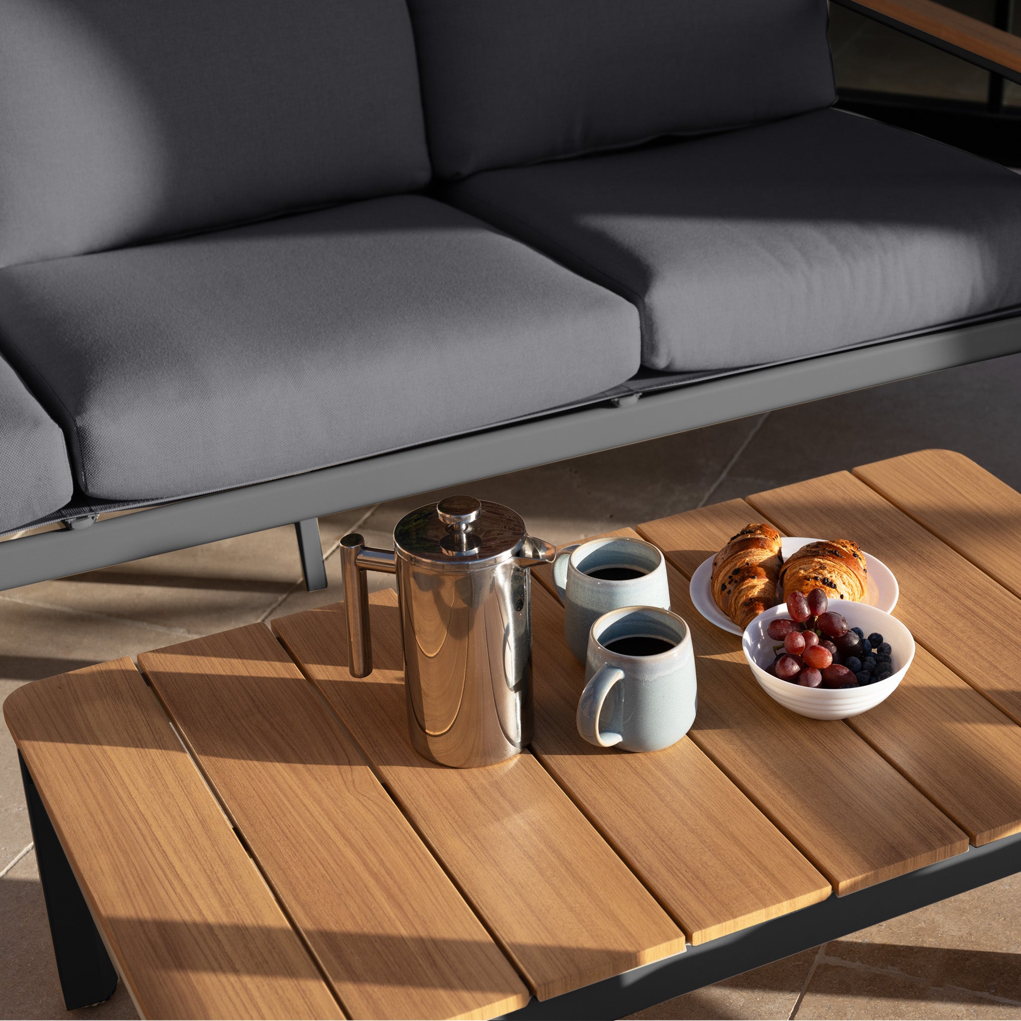 Coffee, croissants, and fruit on a wooden table in front of the Cora 3 Seat Sofa Set in Grey—a modern outdoor sofa set that's weather-resistant and perfect for stylish, comfortable lounging.