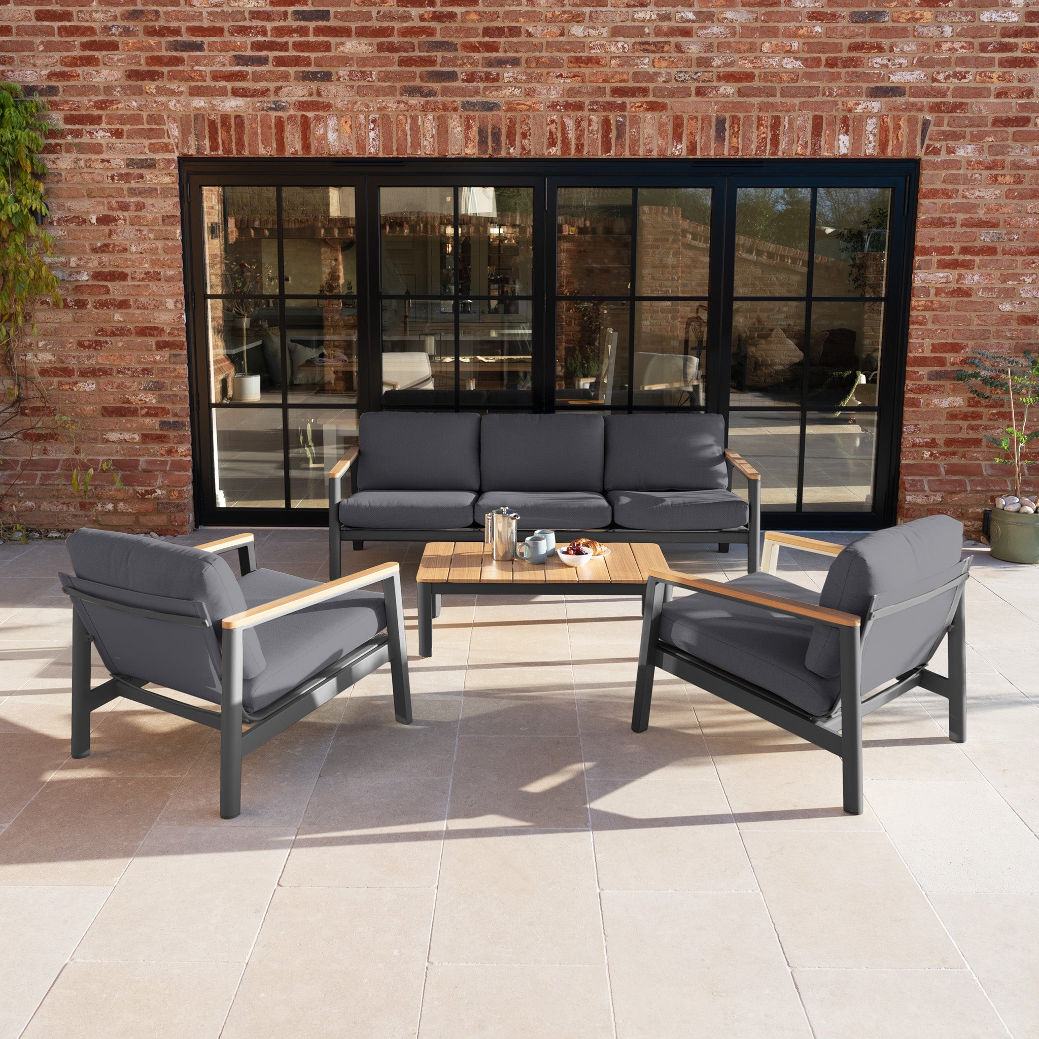 The Cora 3 Seat Sofa Set in Grey adds style to your tiled terrace. This weather-resistant outdoor furniture creates a chic, modern look when placed in front of large glass doors and a brick wall.
