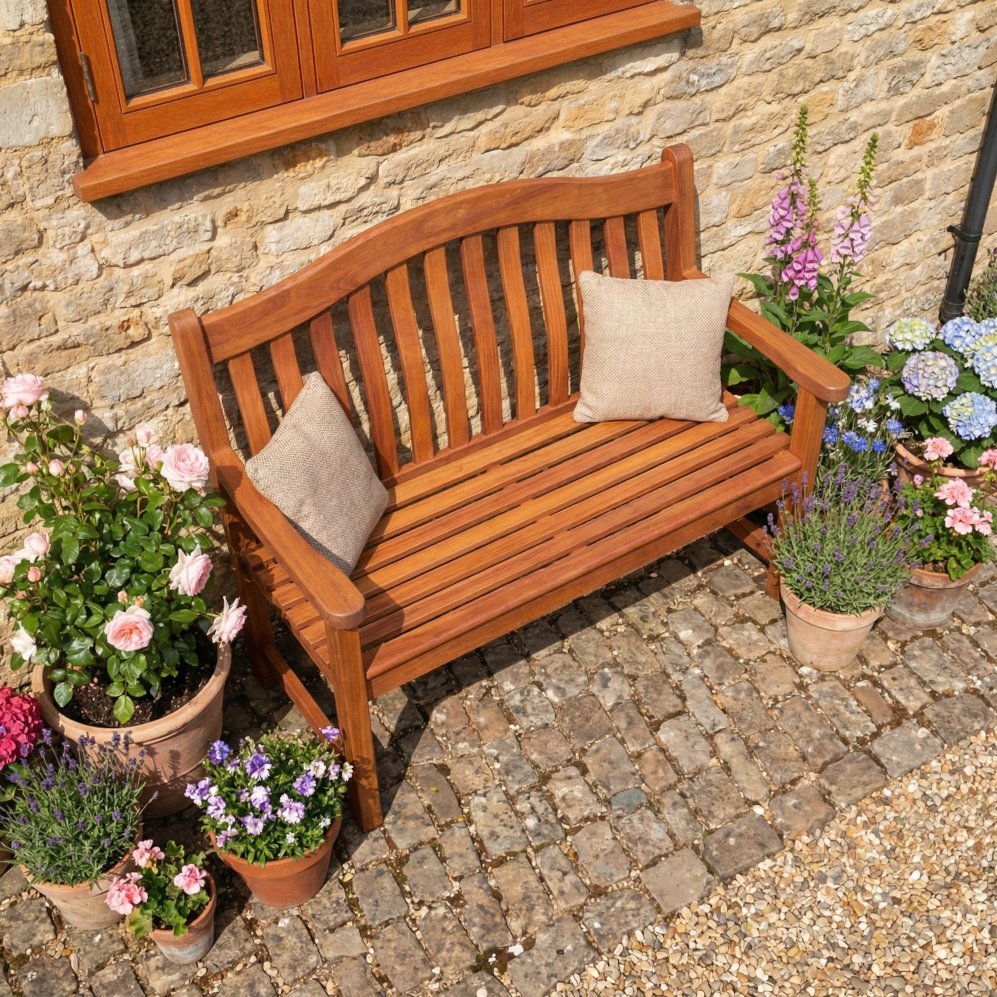 The Cornis Turnberry Bench - 5ft, featuring two cushions, is placed on a cobblestone patio by a brick wall and surrounded by potted flowers.
