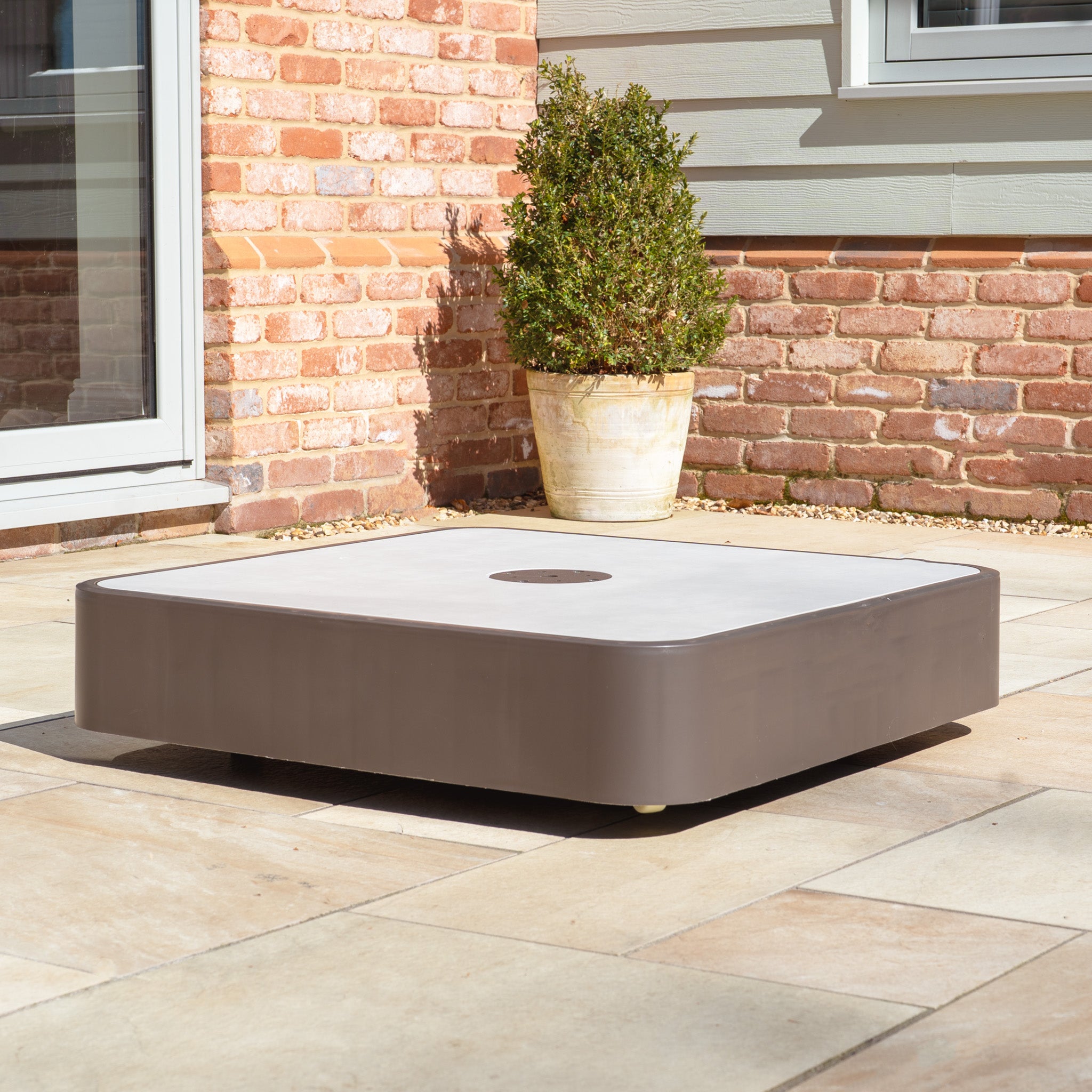 The Costa Fillable Parasol Base with Ceramic Plate sits on the patio, nestled beside a potted plant and brick wall, creating the perfect outdoor retreat.