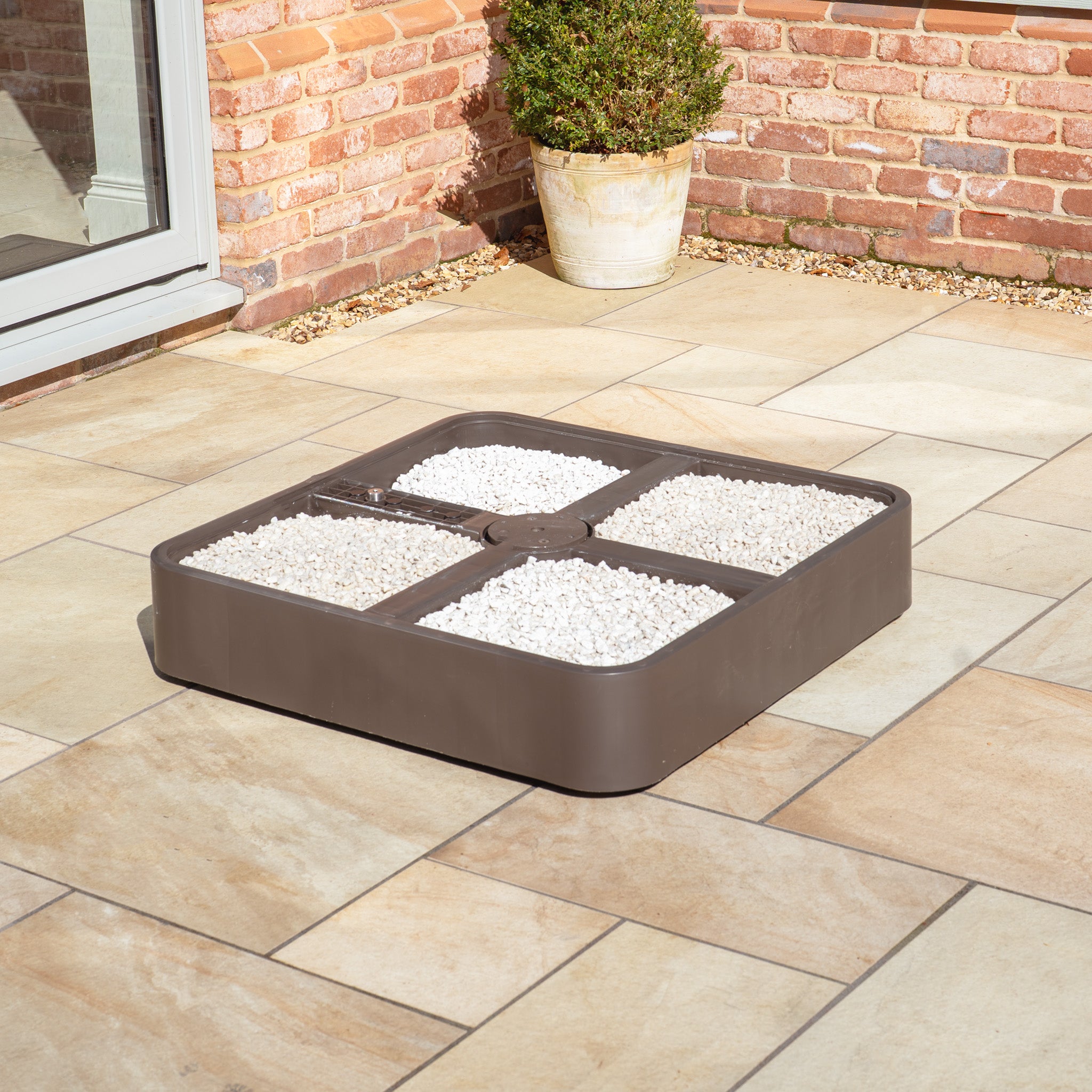 The Costa Fillable Parasol Base with Ceramic Plate ensures stability with its brown square design featuring four sections filled with white pebbles on a patio.
