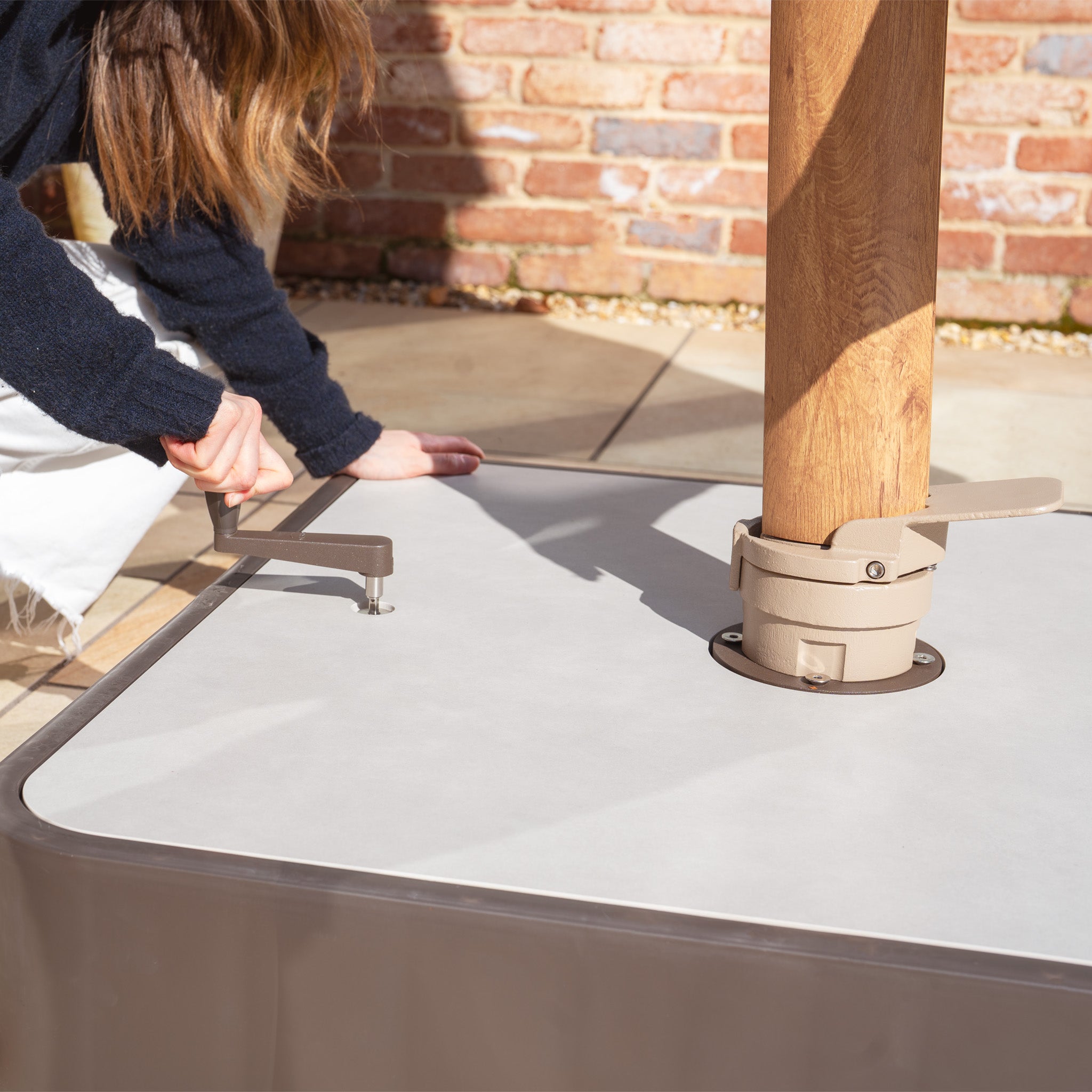 Someone is carefully assembling the Costa Fillable Parasol Base with Ceramic Plate on a tiled patio, with a brick wall behind. Its ceramic cover ensures stability and beautifully enhances any outdoor space.