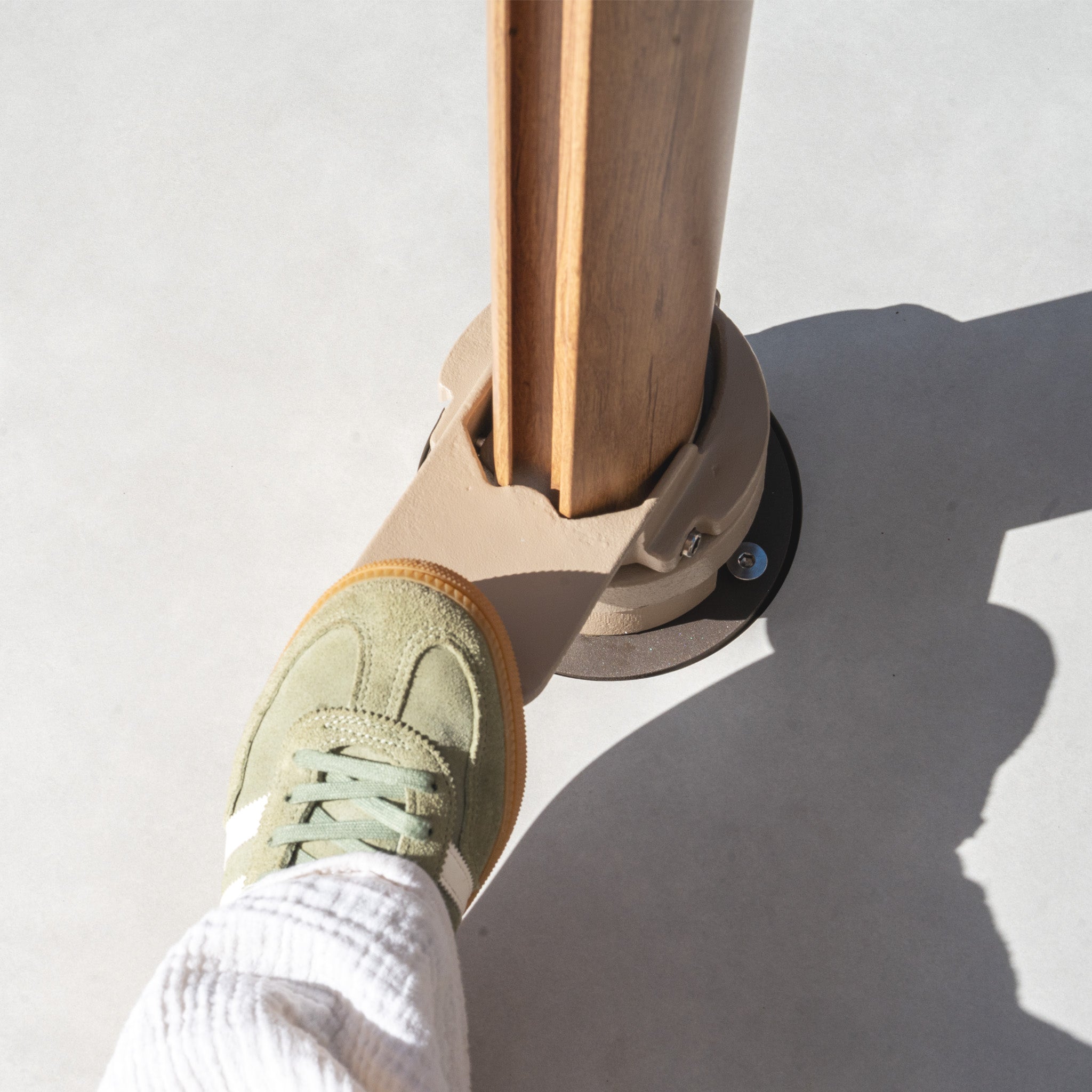 A foot in a green sneaker presses a beige lever on the Costa Fillable Parasol Base with Ceramic Plate against a gray surface, ensuring unwavering stability.