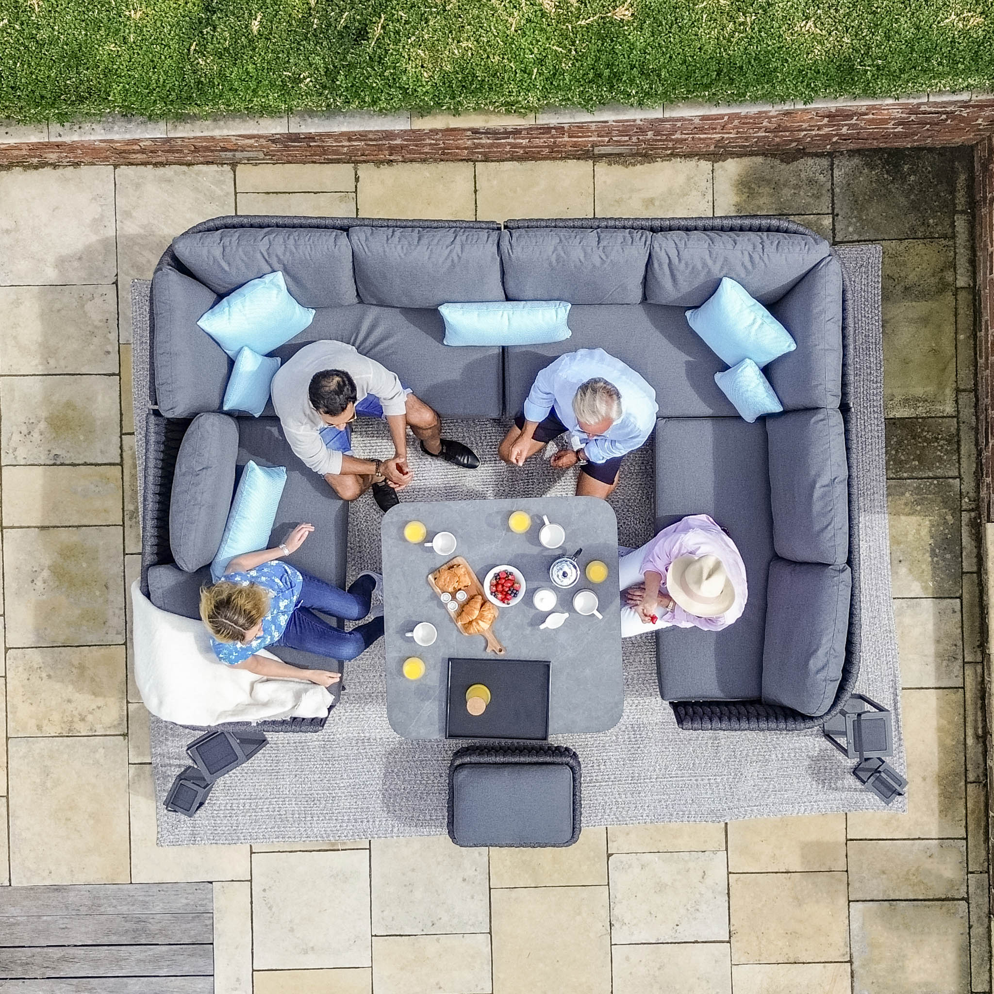 Aerial view of four people sitting on a gray outdoor sectional, part of the Palma U-Shape Rope Corner Dining Set with Rising Table in Grey, surrounded by a patio.