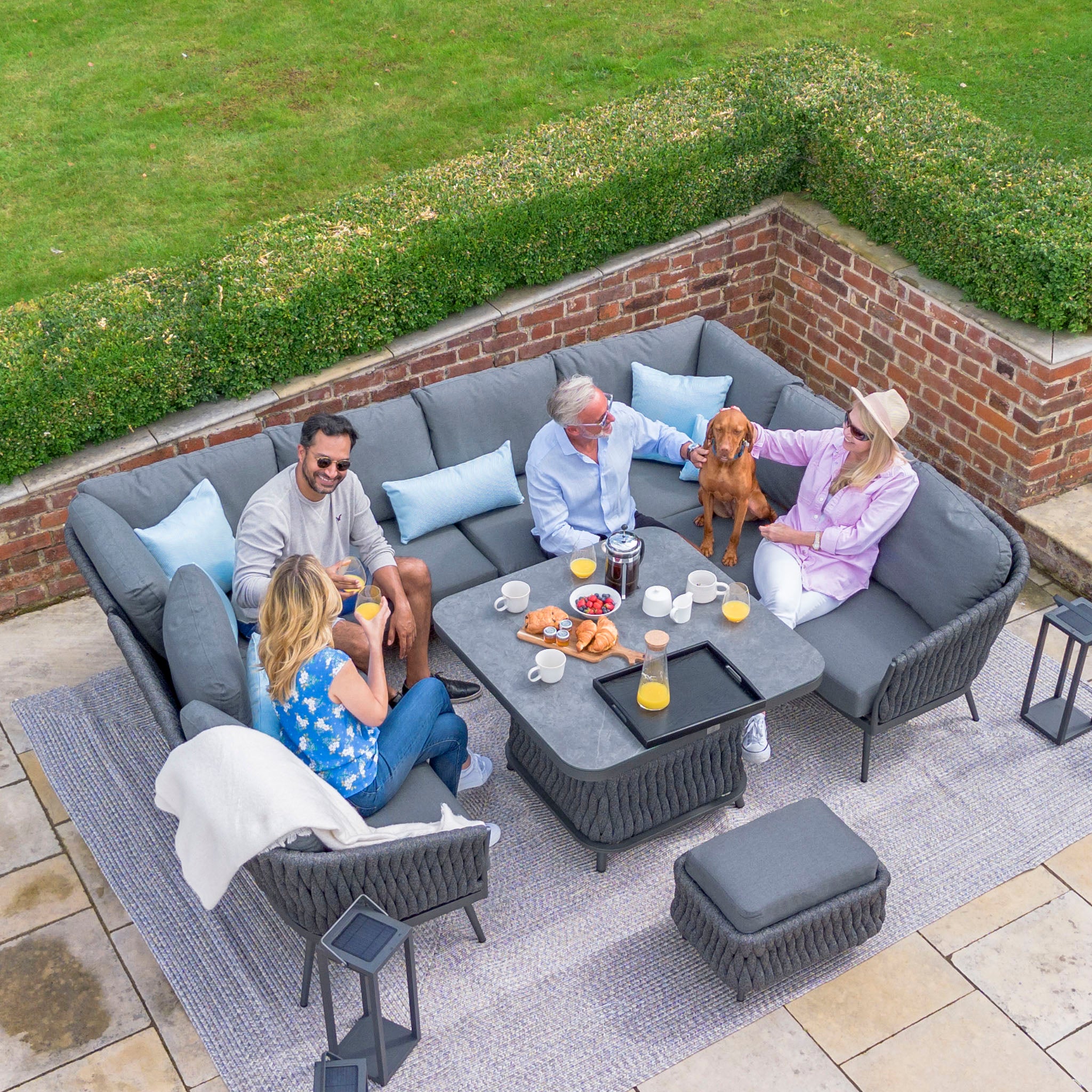 Four people relaxing on a Palma U-Shape Rope Corner Dining Set with Rising Table in Grey, enjoying food and drinks, with one person petting a dog on their lap, all surrounded by the UV resistant fabric that ensures comfort and durability.
