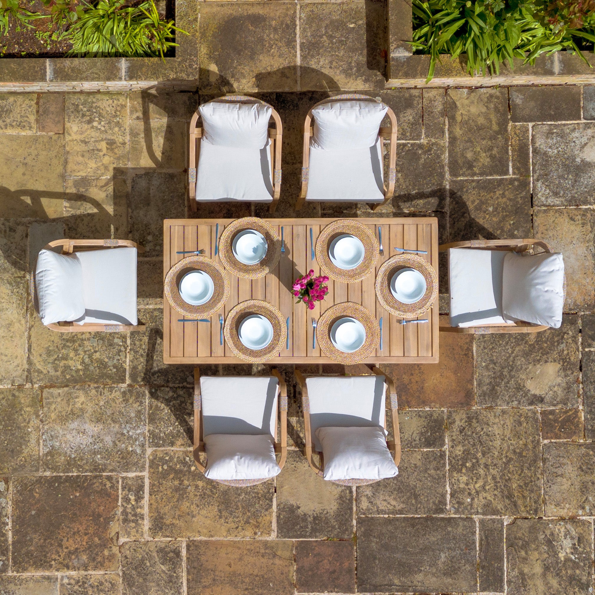 The Quay 6 Seat Rectangular Dining Set in Linen features six cushioned chairs with water-resistant fabric, arranged around a wooden table for six. It is beautifully decorated with a vase of pink flowers and crafted from sustainably sourced acacia wood.