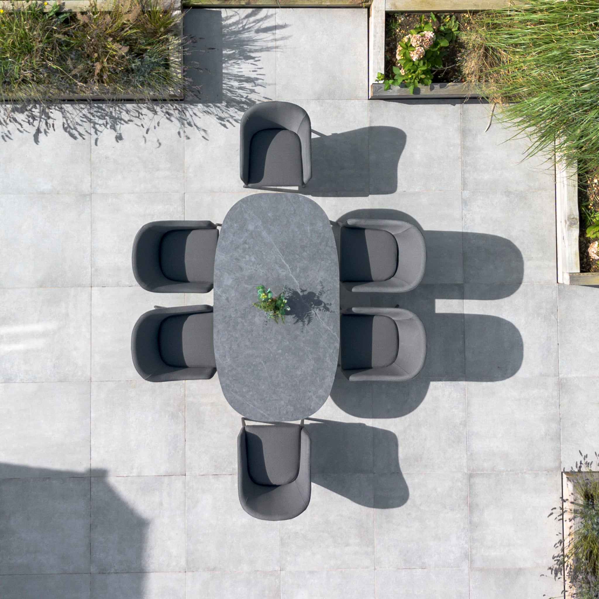 Outdoor dining setup with the Luna 6 Seat Outdoor Fabric Oval Ceramic Dining Set in Grey, featuring six durable gray chairs and a stone table with a small green plant in the center, on a tiled patio.