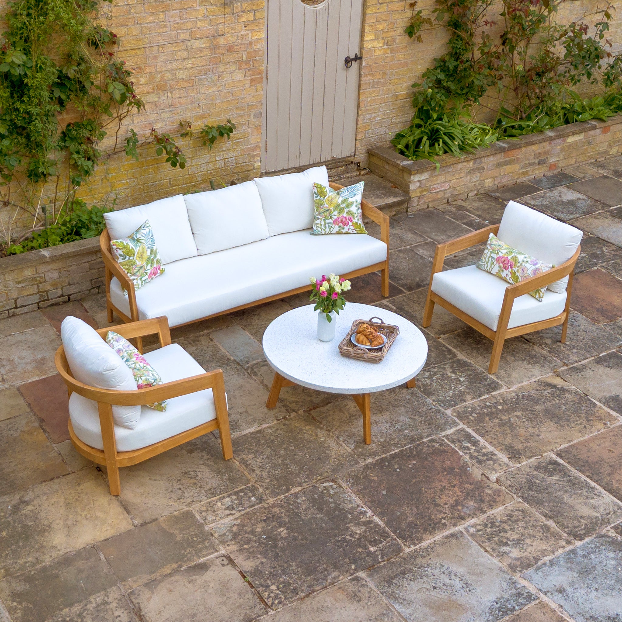 The Yosemite 3 Seat Sofa Set In Glacier includes a white couch, two chairs, and a round table adorned with flowers and snacks, all crafted from premium sustainably sourced teak wood and elegantly displayed on a stone-tiled floor.