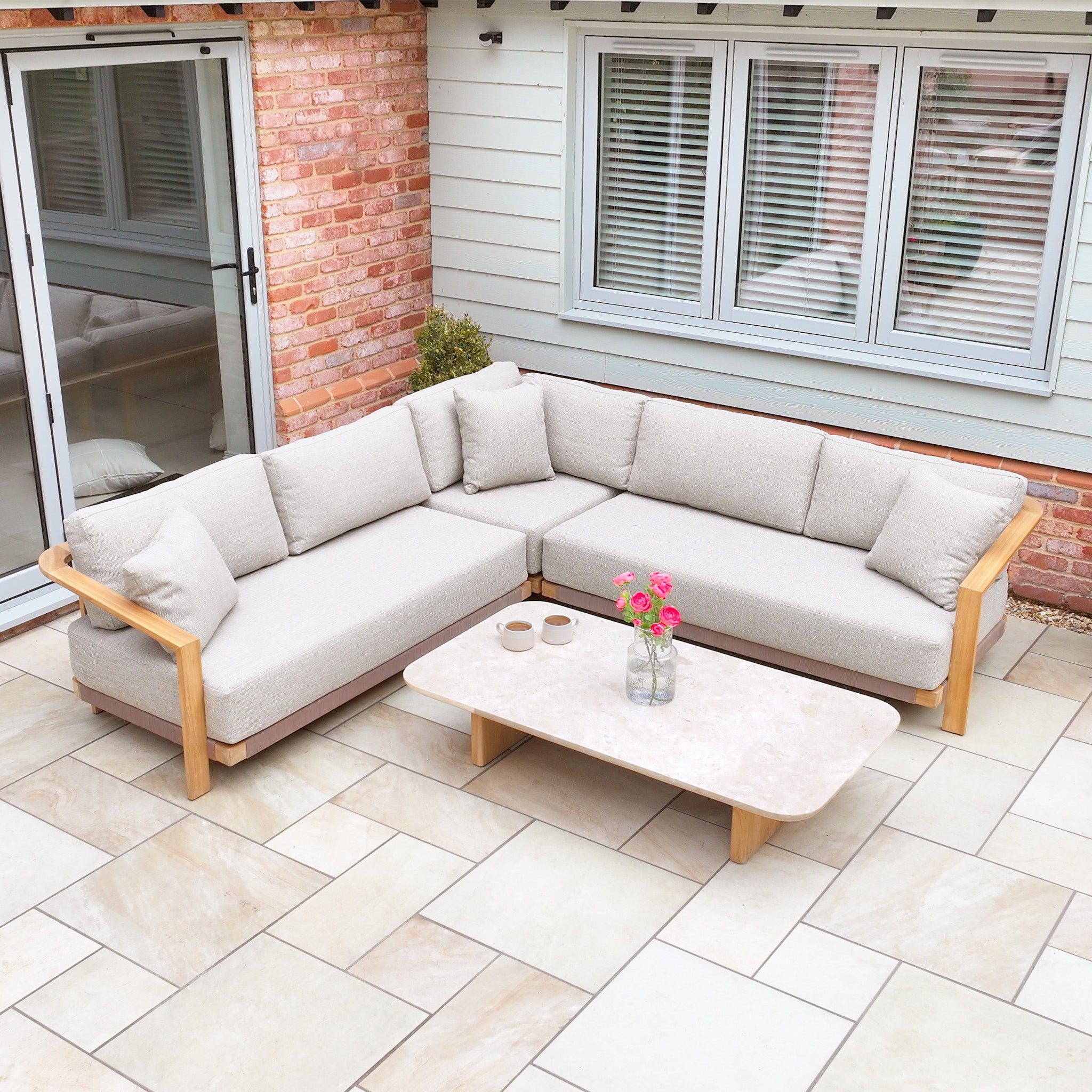 The Tivoli Corner Set with Marble Coffee Table in Fawn is featured on a tiled patio near a brick and wood house, complemented by flowers.
