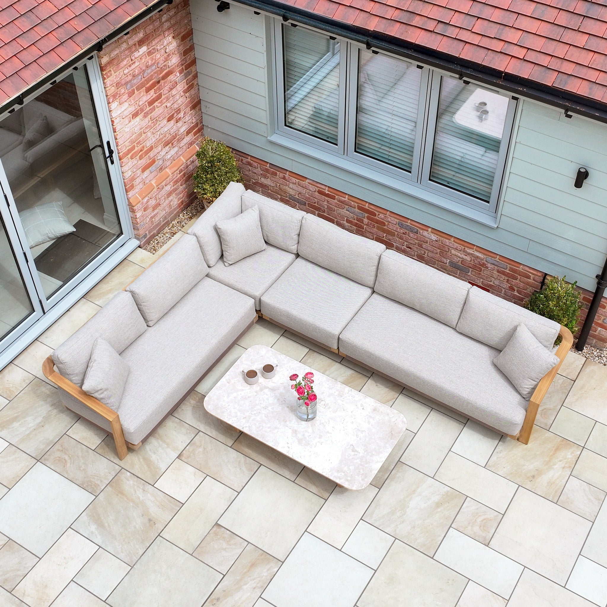 Outdoor patio featuring the Tivoli Large Corner Set with Marble Coffee Table in Fawn and flowers, beside a house with large windows.