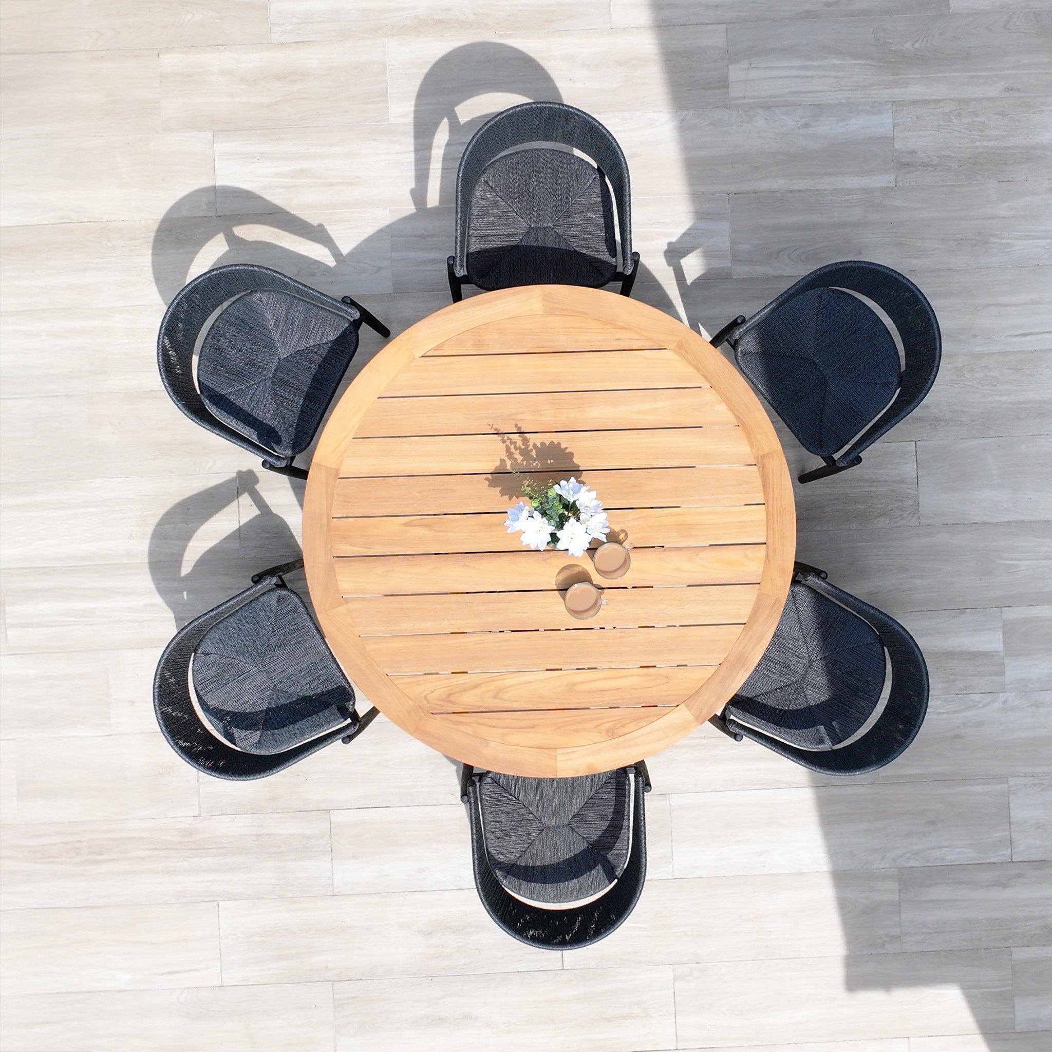 The Lucca 6 Seat Dining Set with Teak Round Table in Black features six black chairs, a teak round table, cups, and a flower vase on a light wooden floor—perfect for stylish outdoor dining on any occasion.