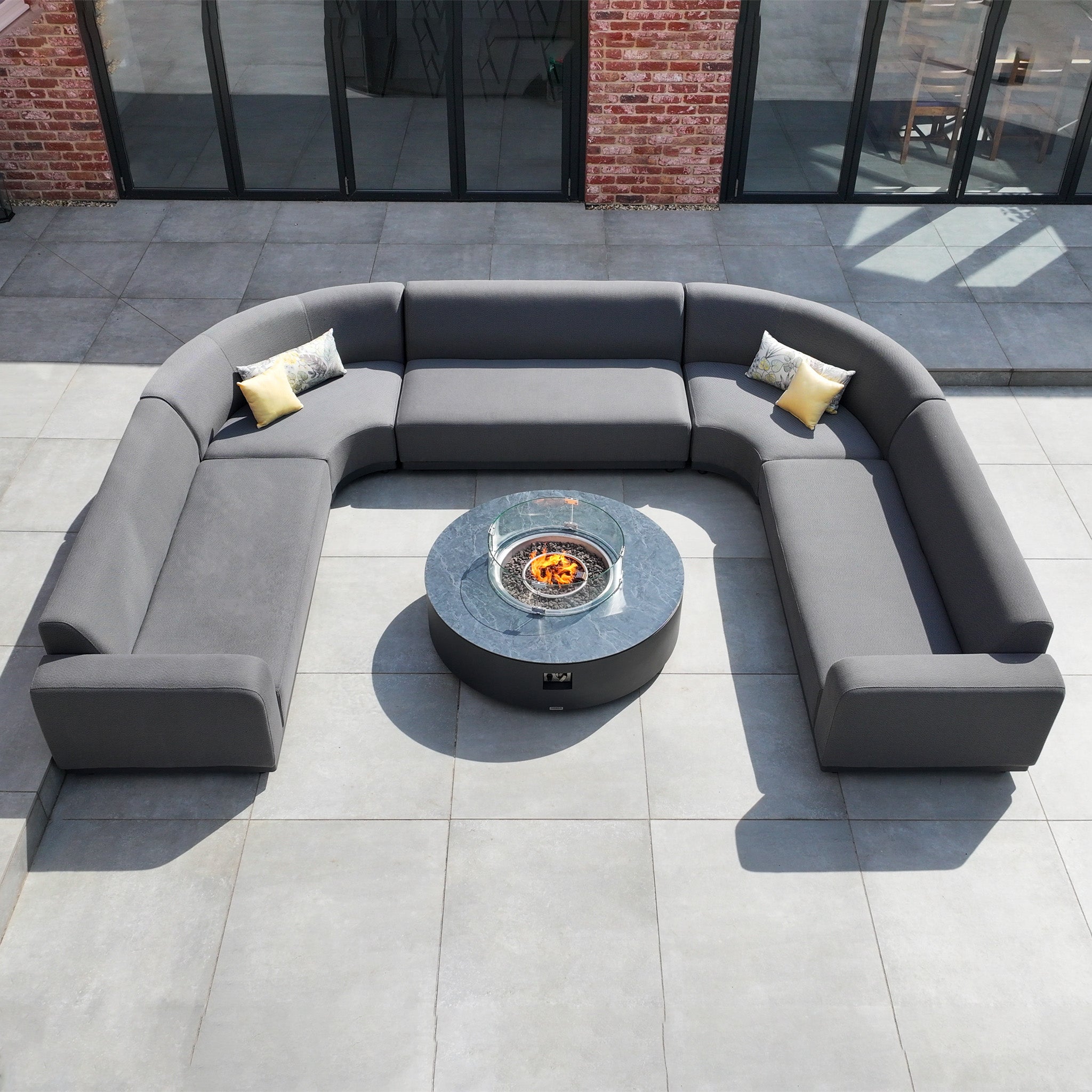 The Luna U-Shape Outdoor Fabric Curved Corner Dining set in grey features a round firepit coffee table and includes three decorative pillows, creating a luxurious outdoor setting on your patio.