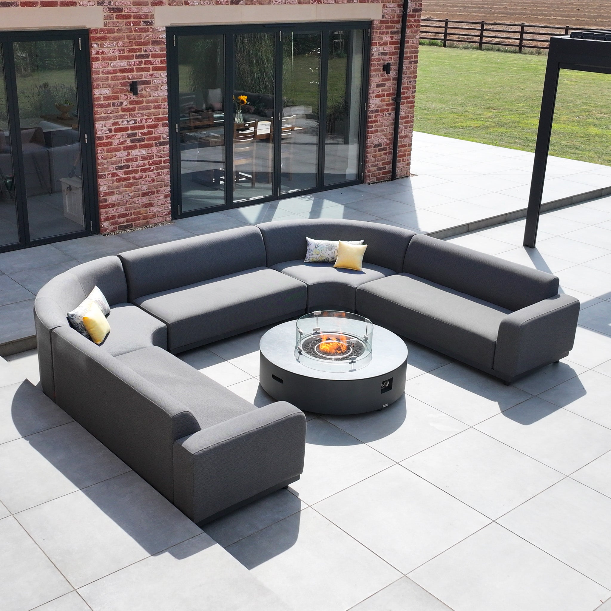 The Luna U-Shape Outdoor Fabric Curved Corner Dining with Round Firepit Coffee Table in Grey creates a stylish and cozy outdoor retreat on a spacious tiled patio by a brick house.