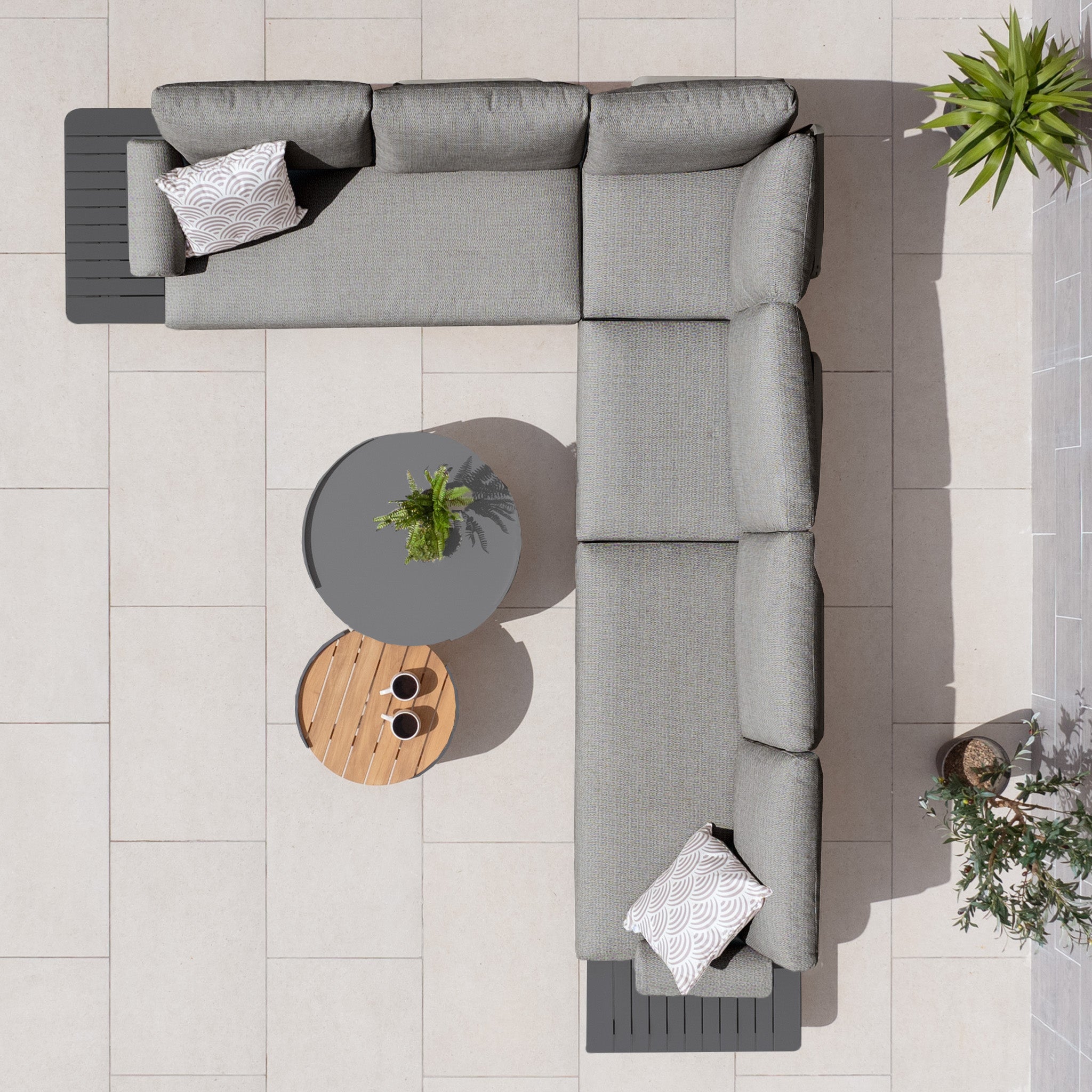 Top view of a Bay Large Corner Group Set with Aluminium Frame in Charcoal, featuring two patterned cushions, an Olefin material finish, and powder-coated aluminum frame, situated next to round tables and potted plants.