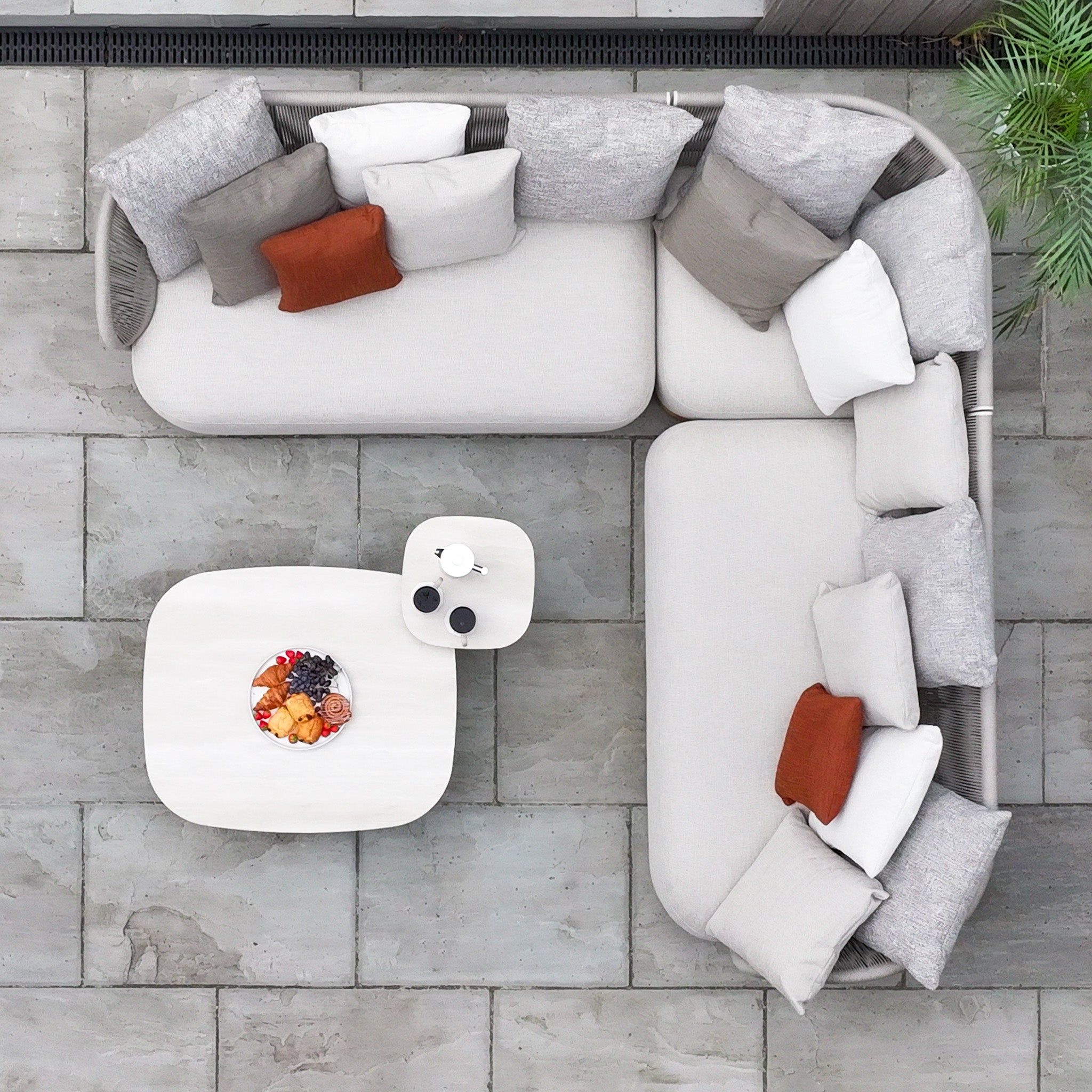 Top view of the Avola Corner Sofa Set in Fawn, a modern outdoor sofa with weather-resistant olefin cushions, two tables, and a fruit platter on a tiled patio.
