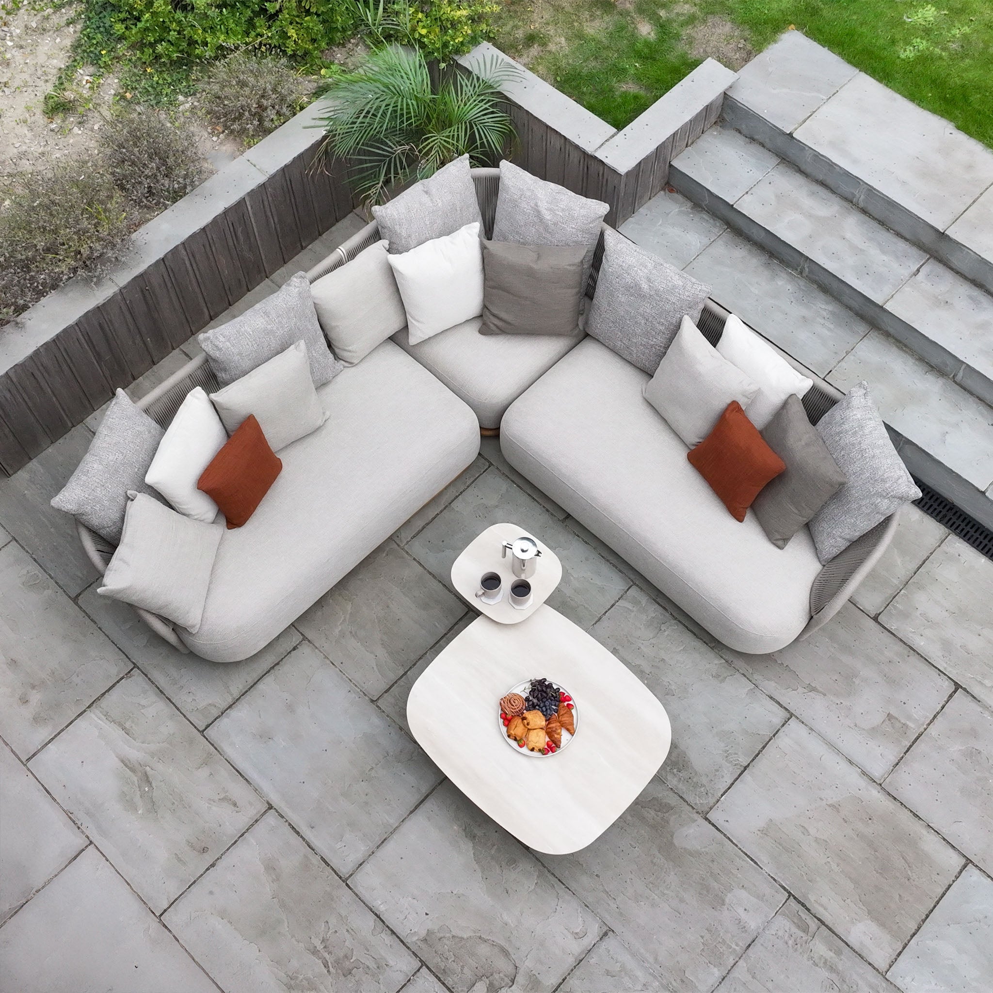 A modern outdoor patio with the Avola Corner Sofa Set in Fawn, showcasing a weather-resistant sofa, plush pillows, and two tables with drinks and snacks, seen from above.