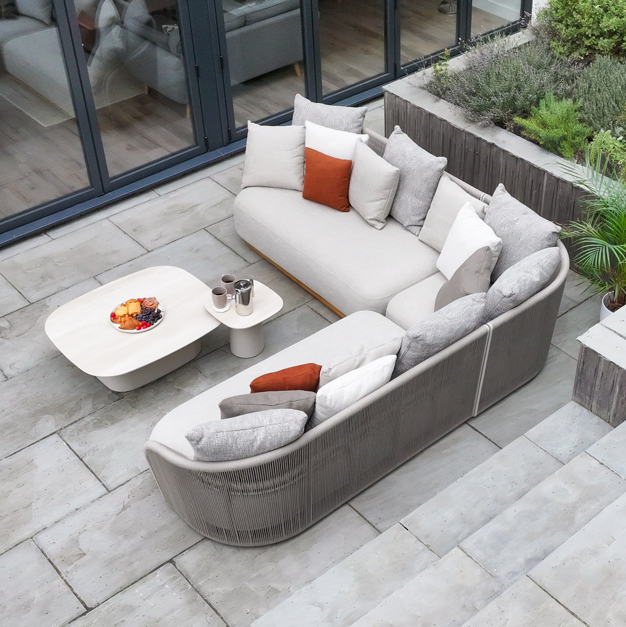 Modern outdoor patio with the Avola Corner Sofa Set in Fawn, featuring weather-resistant olefin cushions, a matching coffee table, and potted plants on stone tiles.