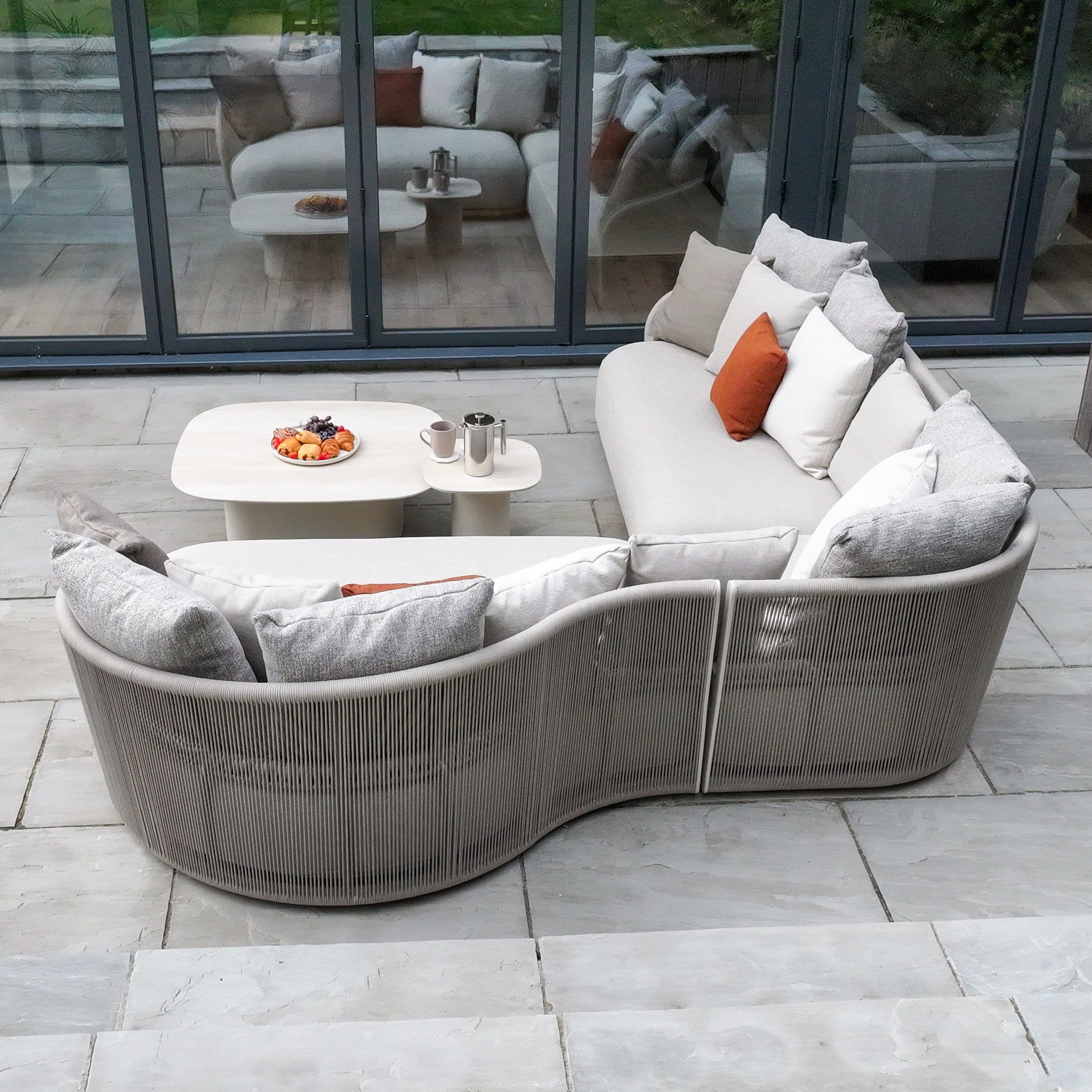 Avola Deluxe Corner Sofa Set in Fawn (Left Hand) with weather-resistant cushions and a white coffee table topped with fruit and mugs, arranged on a stone patio.