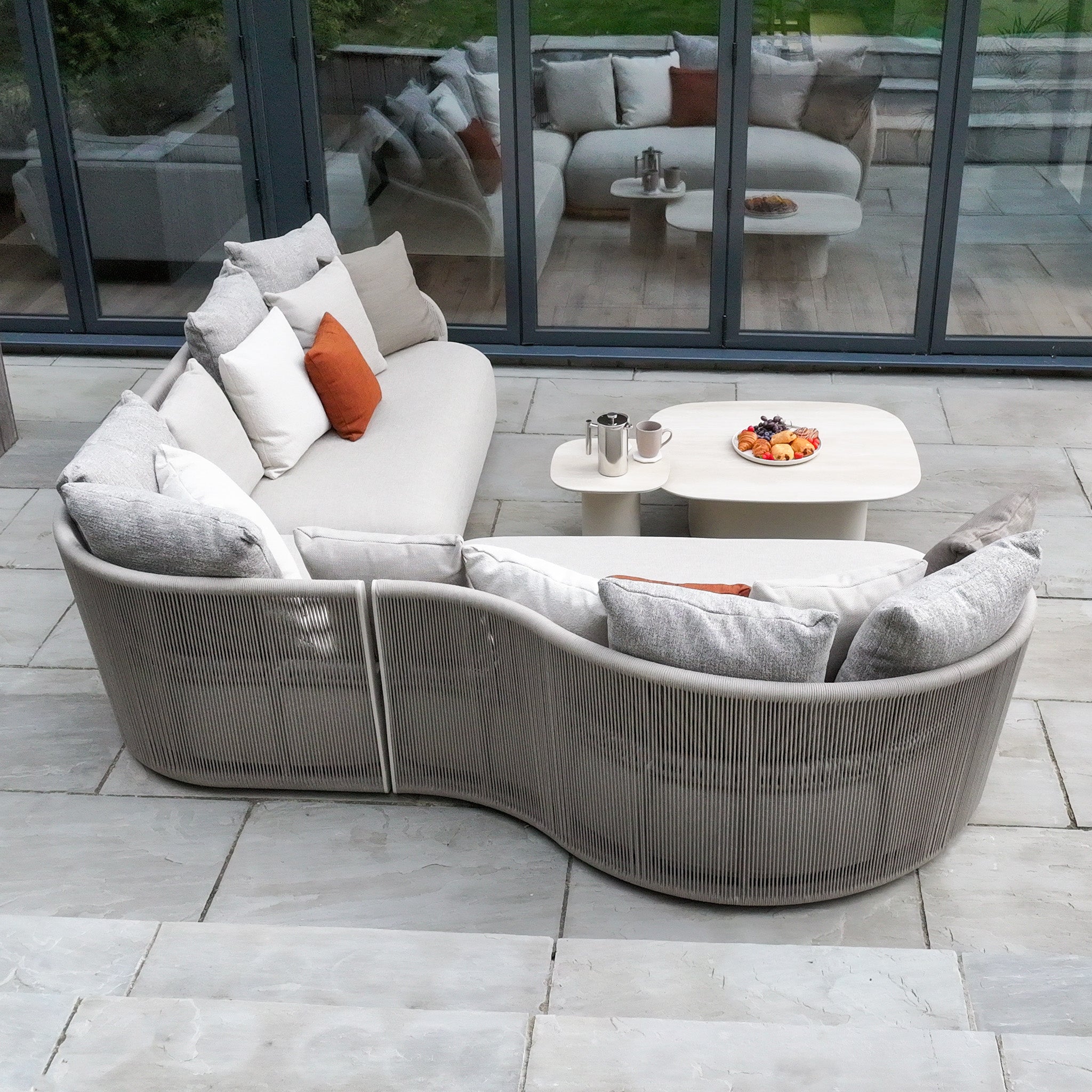 The Avola Deluxe Corner Sofa Set in Fawn (Right Hand) includes a curved outdoor sofa with weather-resistant Olefin rope, comfortable cushions, and a coffee table—perfect for relaxing on a stone patio by glass doors.