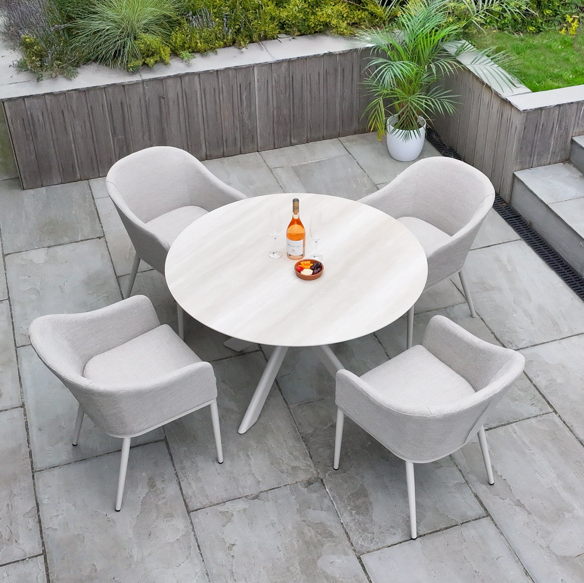 The Luna 4 Seat Round Outdoor Fabric Dining Set with Albatross Table in Fawn features a round table and four beige chairs on a stone patio, accented by a wine bottle, glasses, and bowl—an elegant all-weather set for your outdoor space.