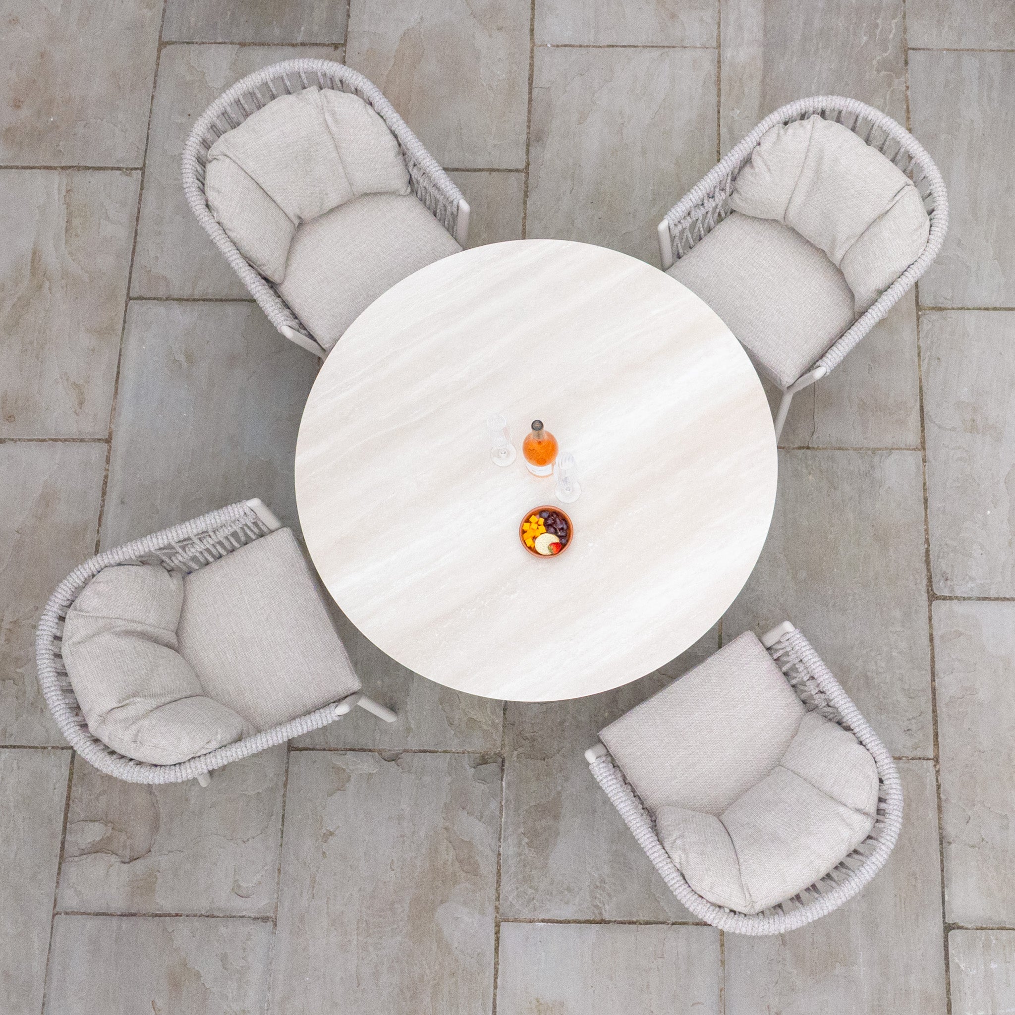 The Pello 4 Seat Round Dining Set with Albatross Table in Fawn is beautifully arranged with four cushioned chairs, orange juice, and a fruit bowl on a stone tile floor—an inviting scene for stylish outdoor dining.