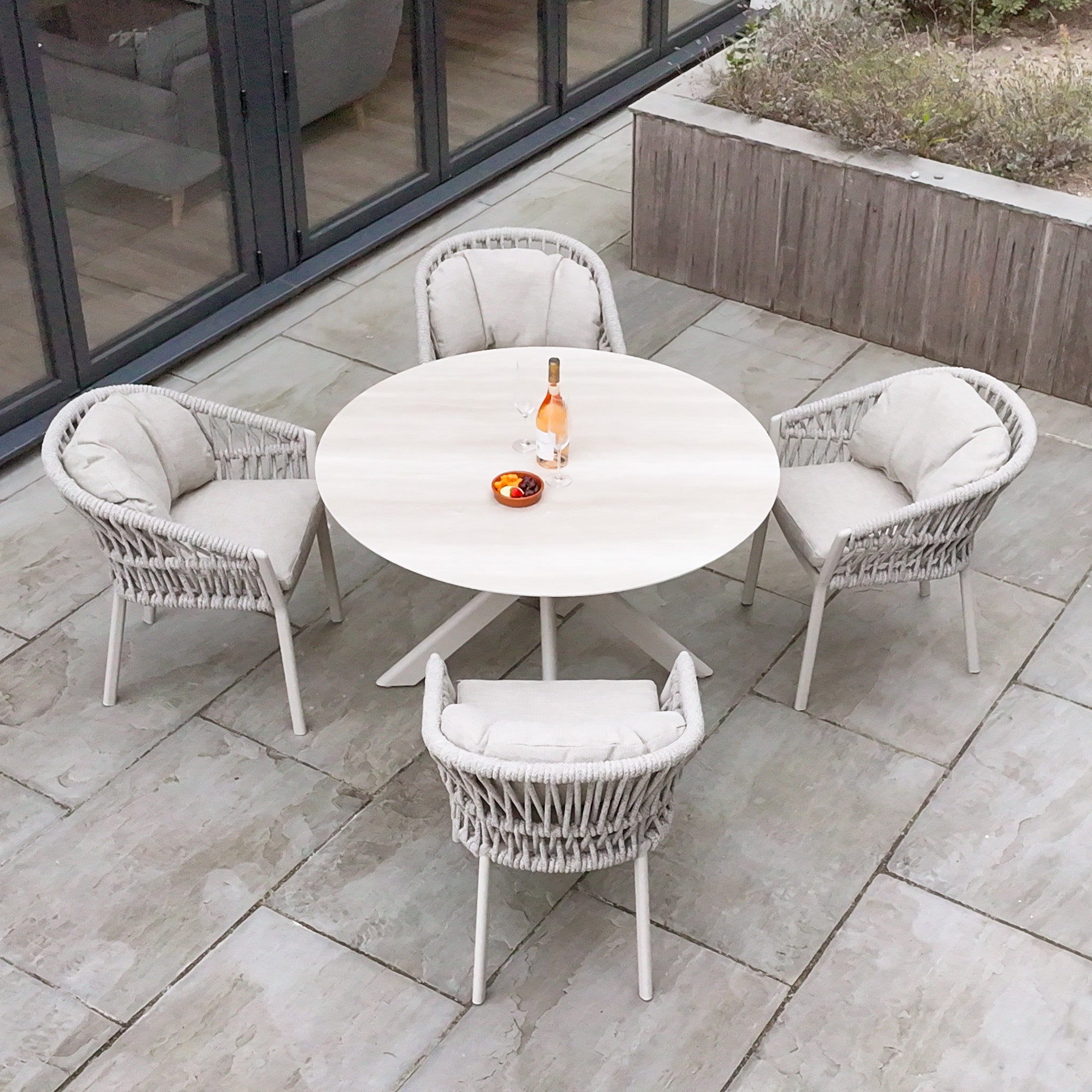 The Pello 4 Seat Round Dining Set with Albatross Table in Fawn, arranged on a tiled patio and styled with wine and fruit, creates the perfect outdoor dining setup.