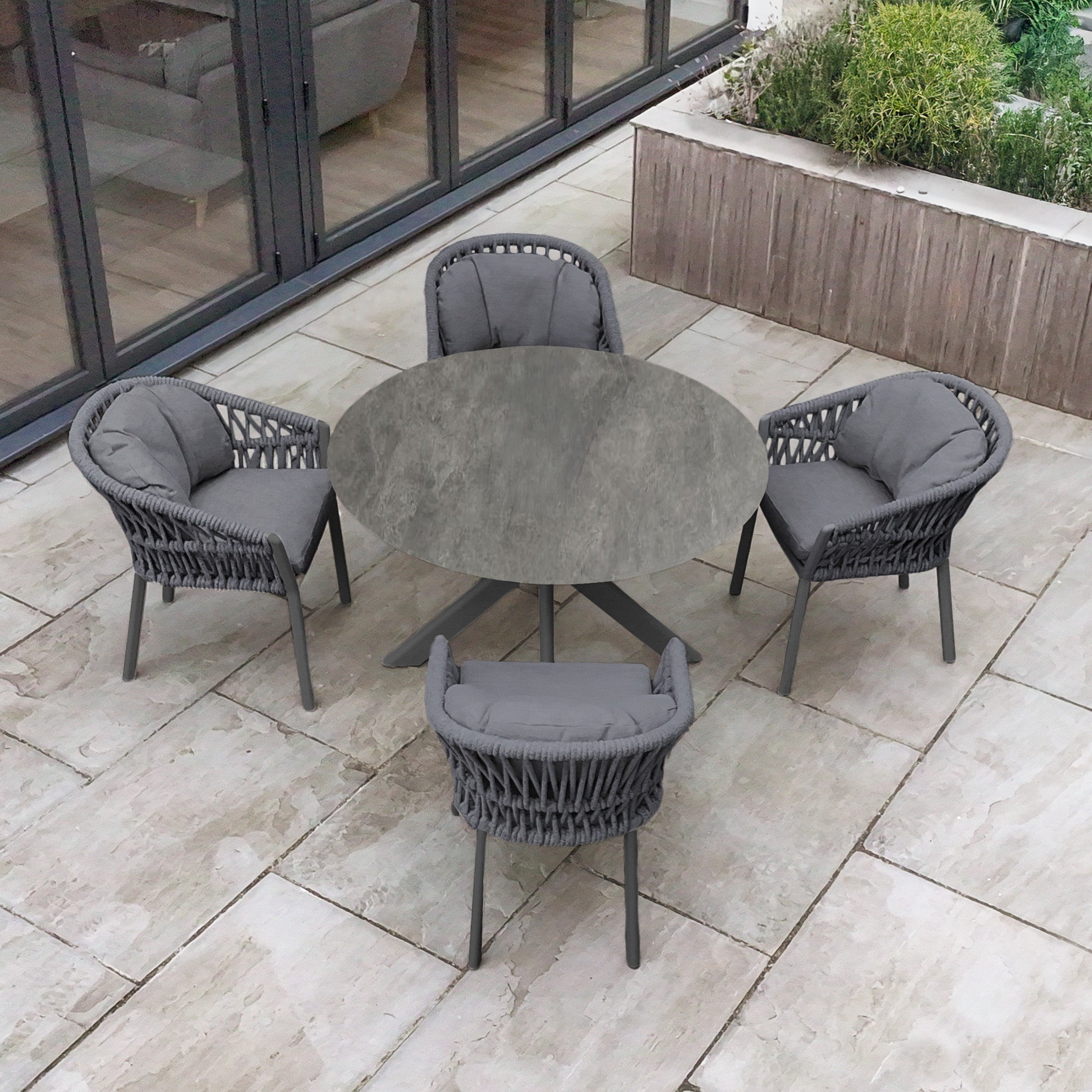 A Pello 4 Seat Round Dining Set with Albatross Table in Grey, featuring cushioned chairs, is arranged on a stone patio beside large windows and a planter box.