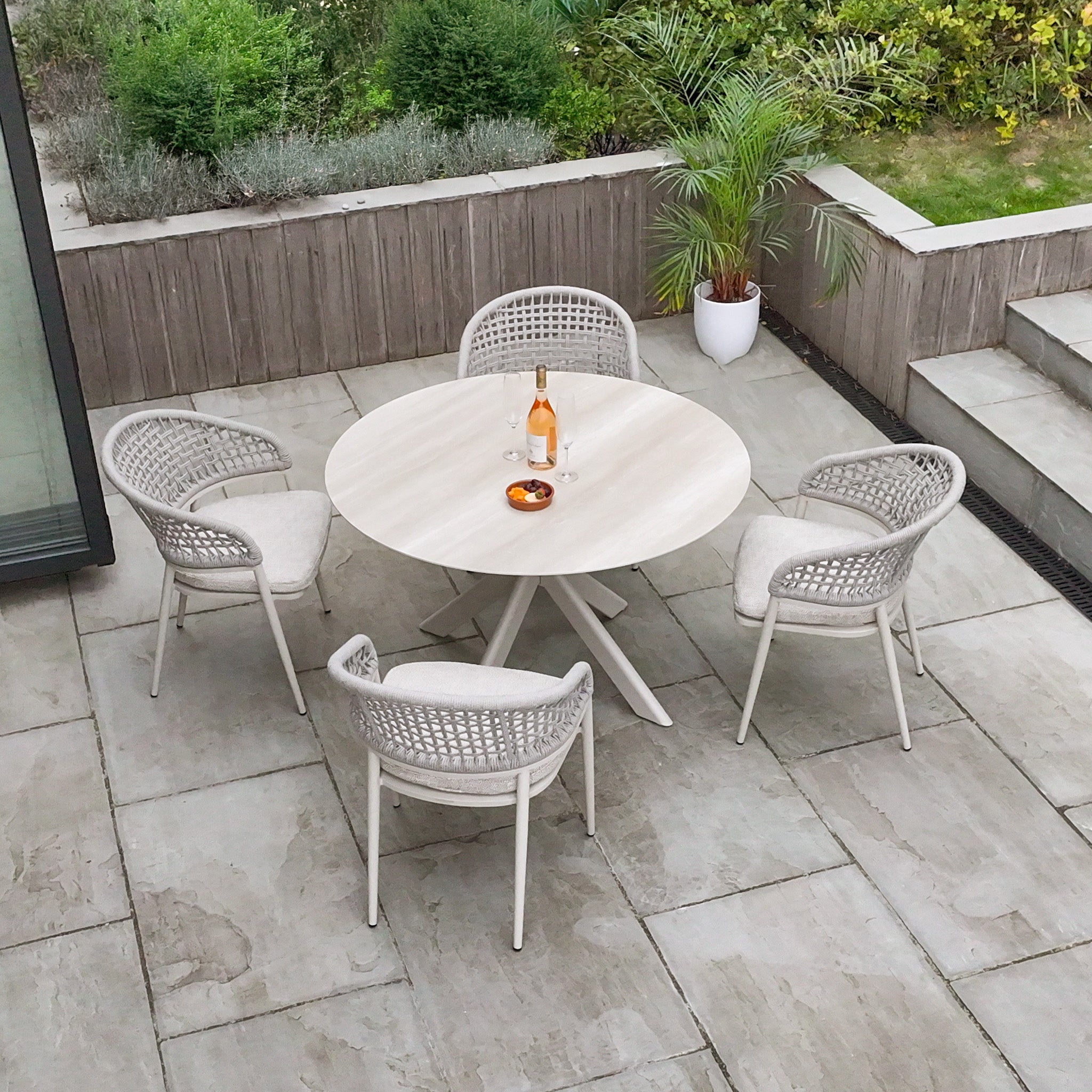 The Kalama 4 Seat Round Rope Dining Set with Albatross Table in Oatmeal creates a cozy patio retreat, featuring a round table and four chairs. A potted plant and drinks on the stylish set complete the inviting garden scene.