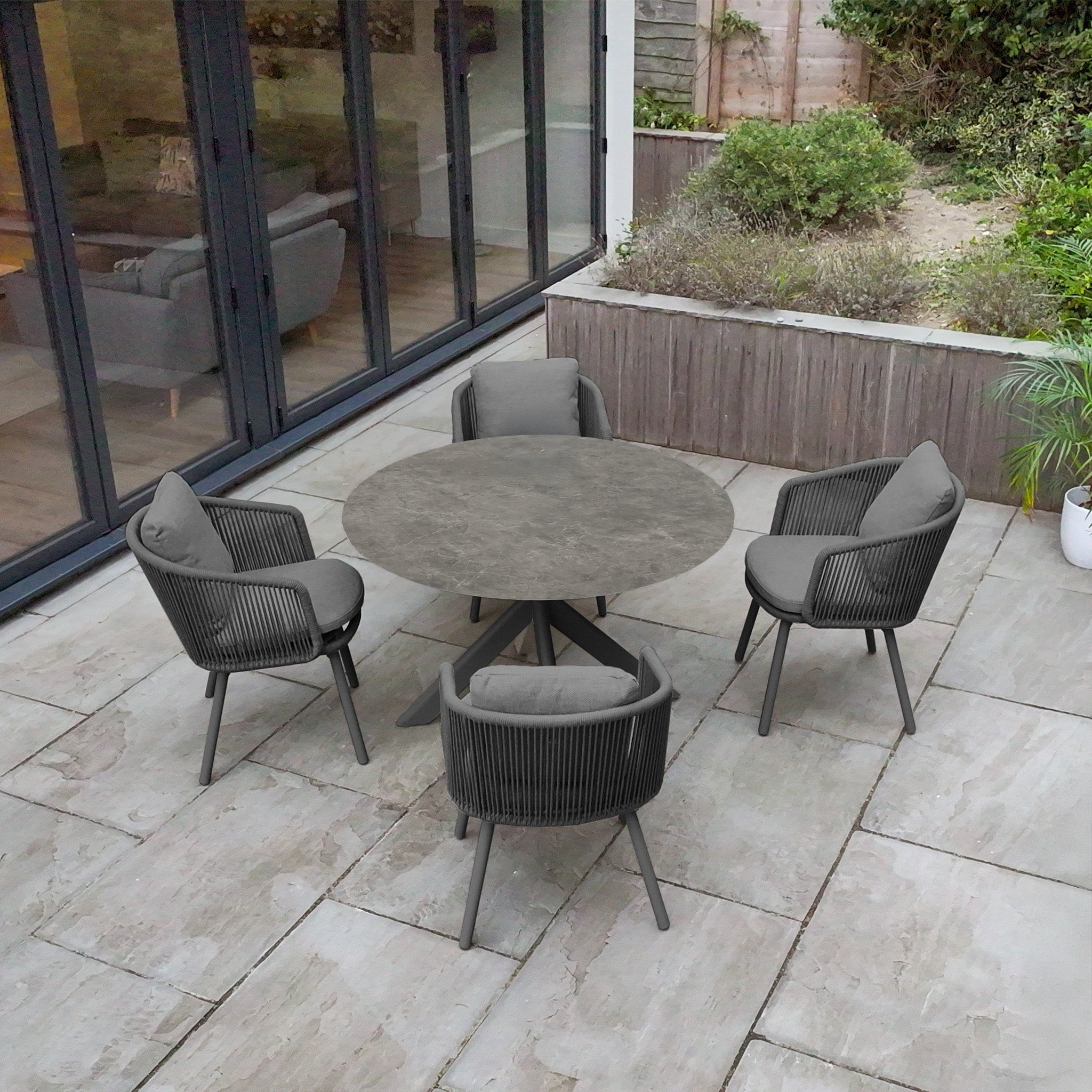 The Dekota 4 Seat Round Rope Dining Set with Albatross Table in Charcoal creates a stylish outdoor dining space on a modern patio beside glass doors and a garden area.
