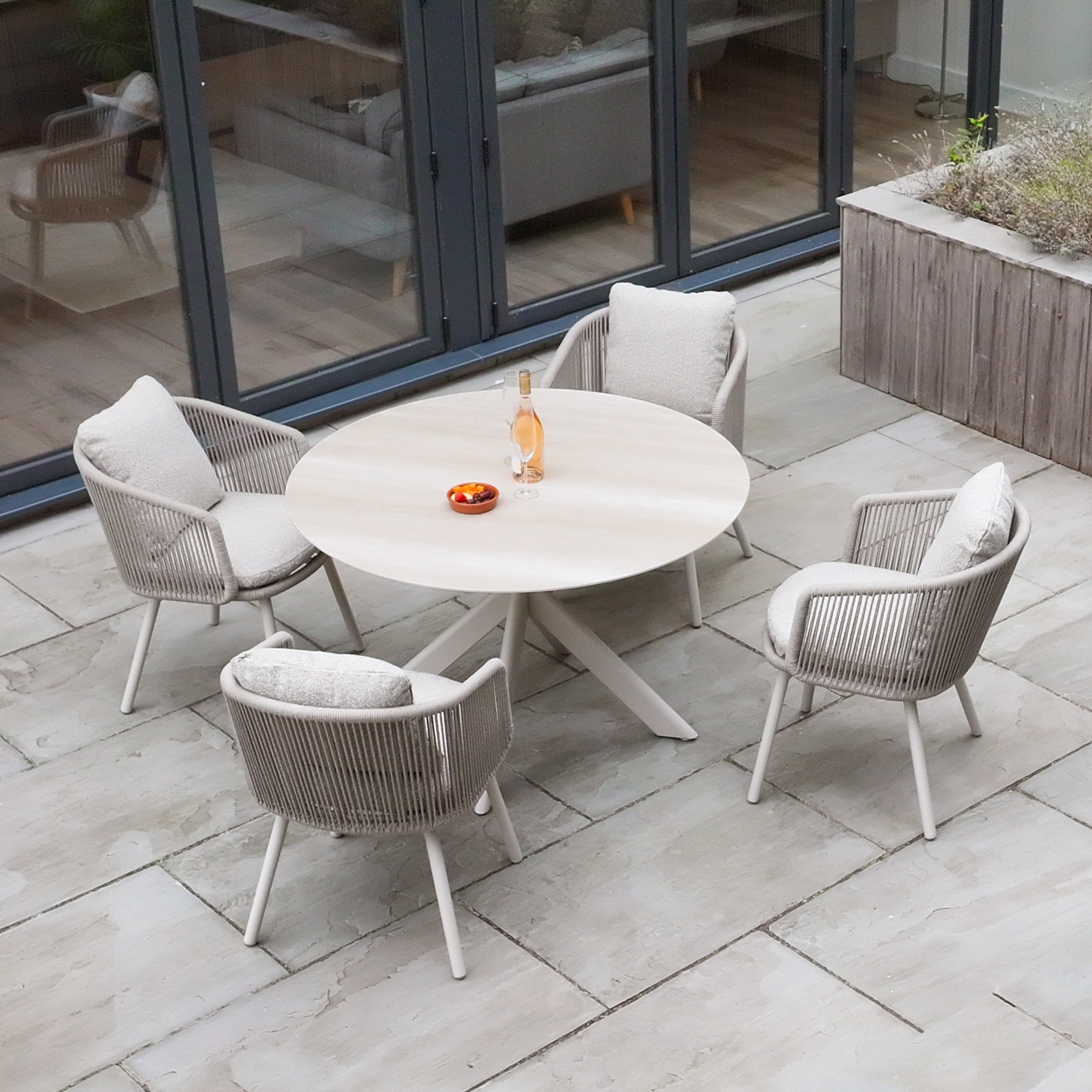 Theia 4 Seat Round Rope Dining Set with Albatross Table in Oatmeal, styled for outdoor dining on a tiled patio, features cushioned chairs and is ideal for garden gatherings.