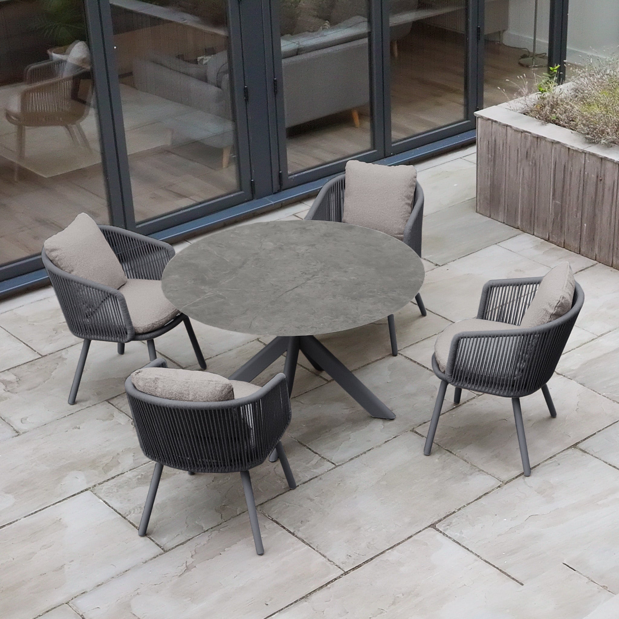The Theia 4 Seat Round Rope Dining Set with Albatross Table in Truffle features four cushioned chairs and a round stone table, creating an inviting and stylish outdoor dining area ideal for gatherings.