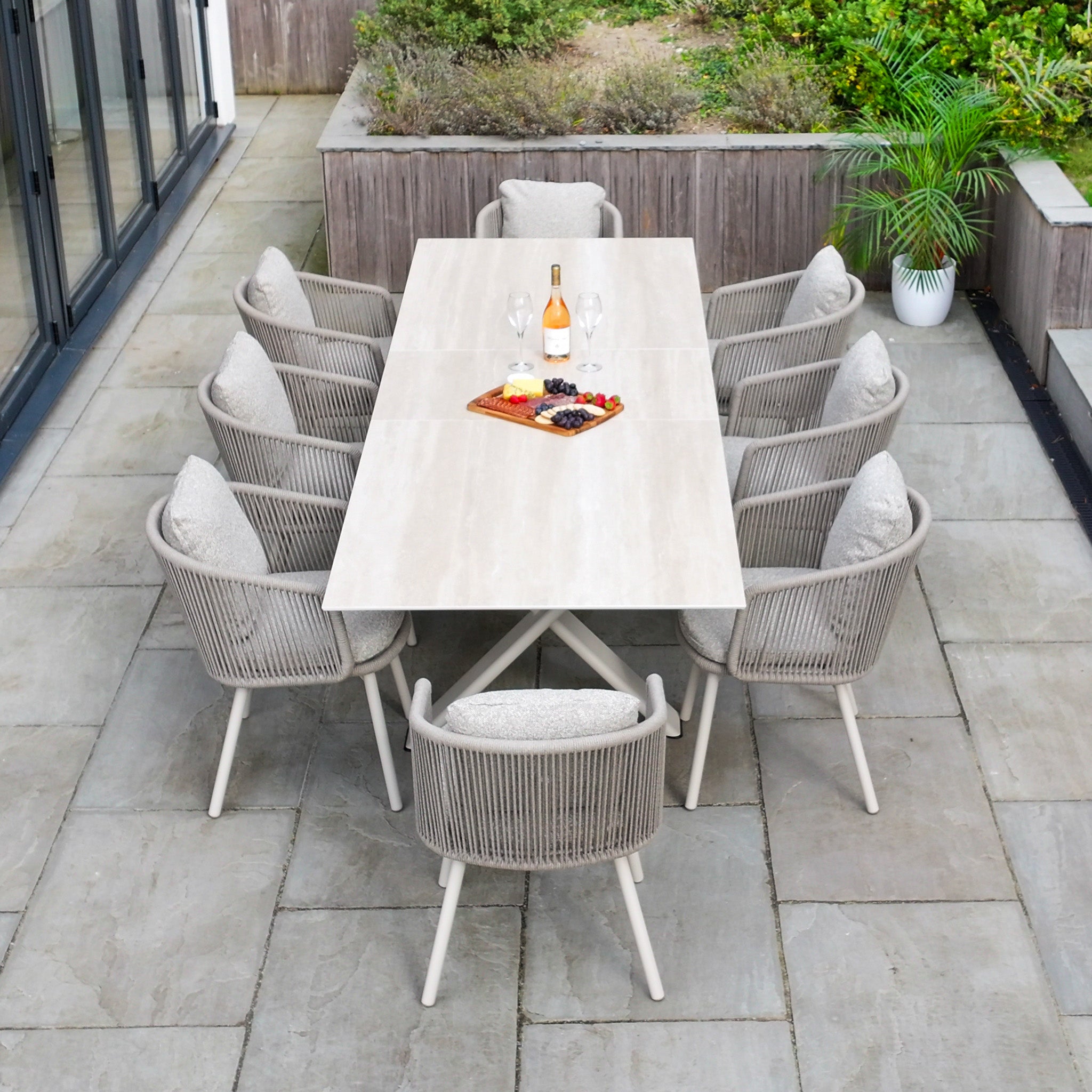 The Theia 8 Seat Rope Dining Set with Albatross Extending Table in Oatmeal is arranged on a stone patio with wine, glasses, and snacks—ideal for outdoor gatherings.