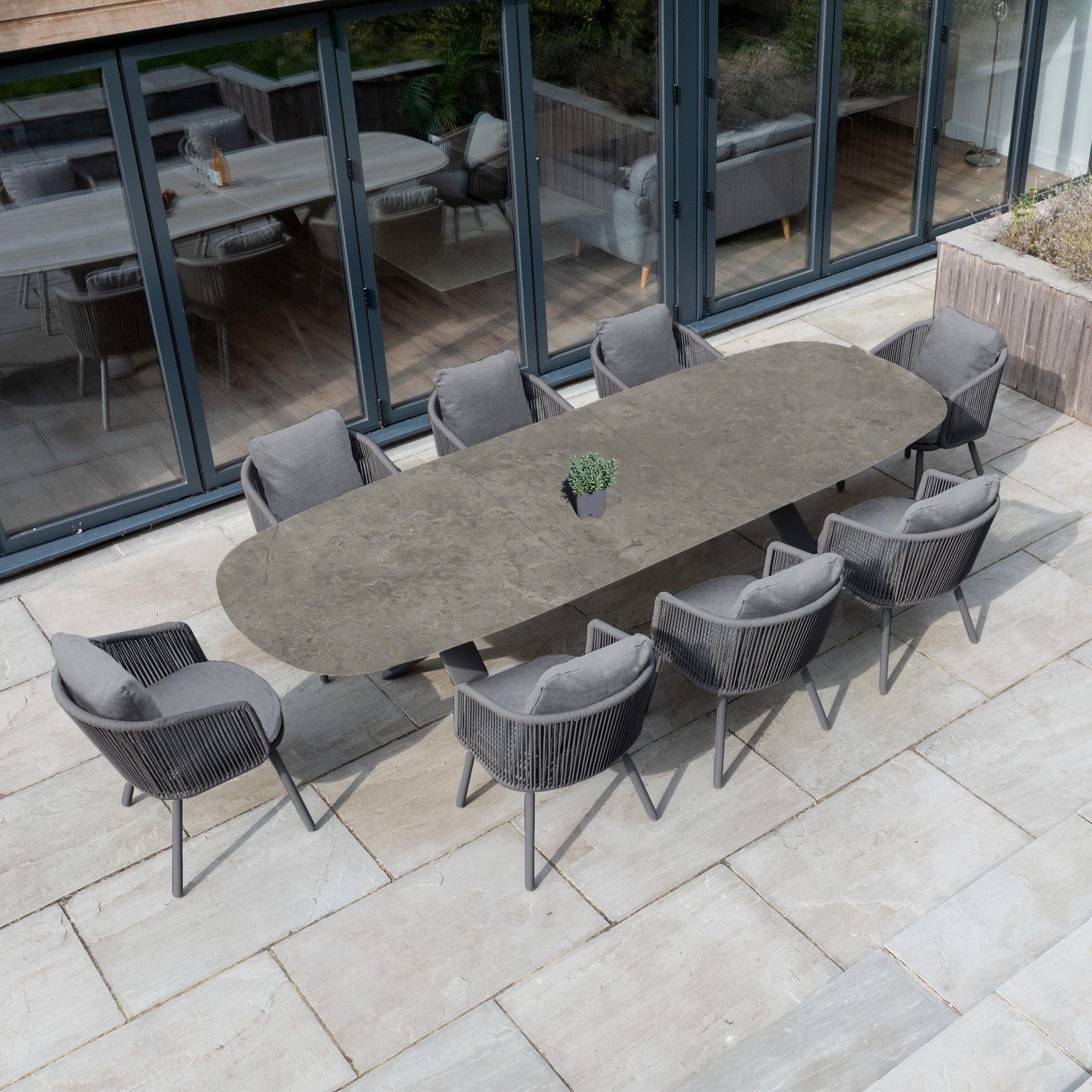 The Dekota 8 Seat Oval Rope Dining Set with Albatross Extending Table in Charcoal includes eight gray cushioned chairs with rope accents, arranged around a table centerpiece on a stone patio.