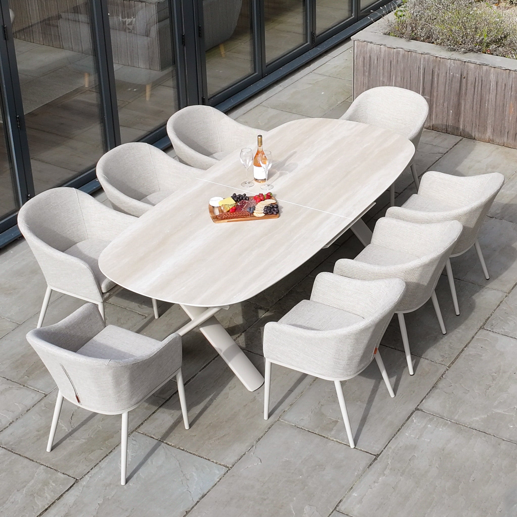 The Luna 8 Seat Outdoor Fabric Oval Dining Set with Albatross Extending Table in Fawn features eight light gray chairs, UV resistant cushions, and is shown on a stone patio with wine and a fruit platter.