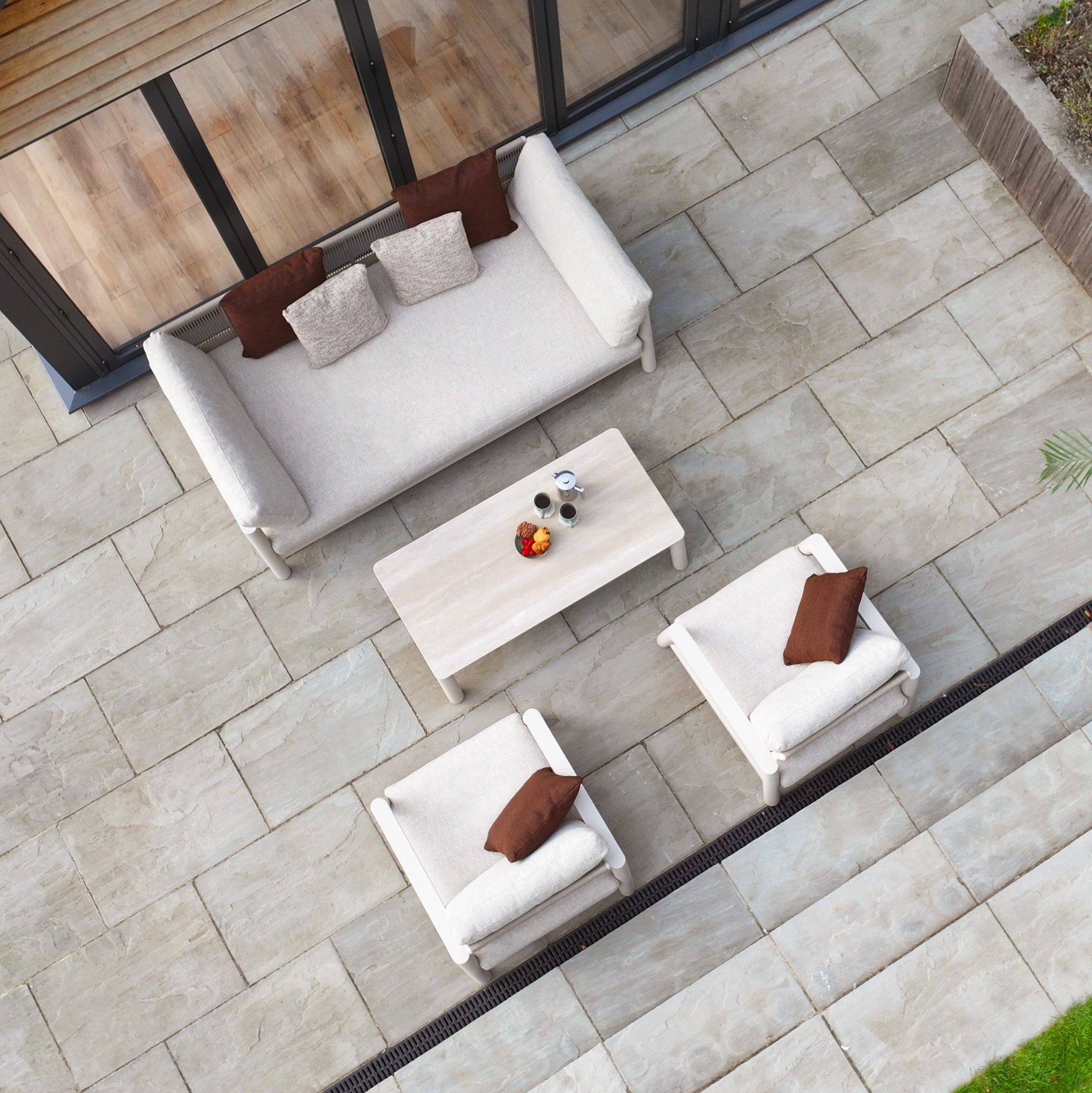 Outdoor patio featuring the Theia 3 Seat Sofa Set in Oatmeal, including a sofa, two chairs, a table with drinks, and neatly arranged beige tiles.