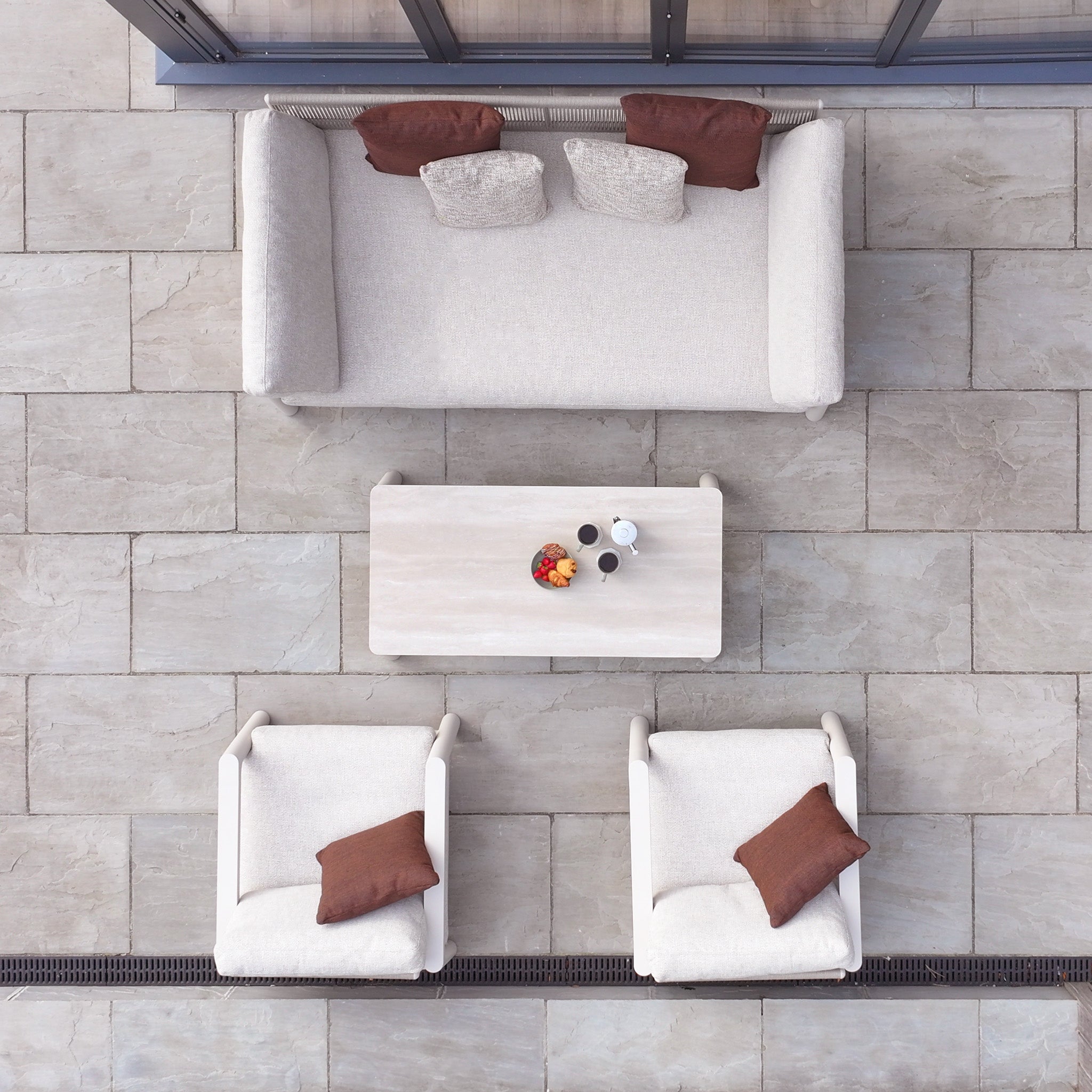 Outdoor patio with Theia 3 Seat Sofa Set in Oatmeal, two chairs, coffee table, brown pillows, and fruit bowl—a perfect spot to enjoy stylish modular garden furniture.