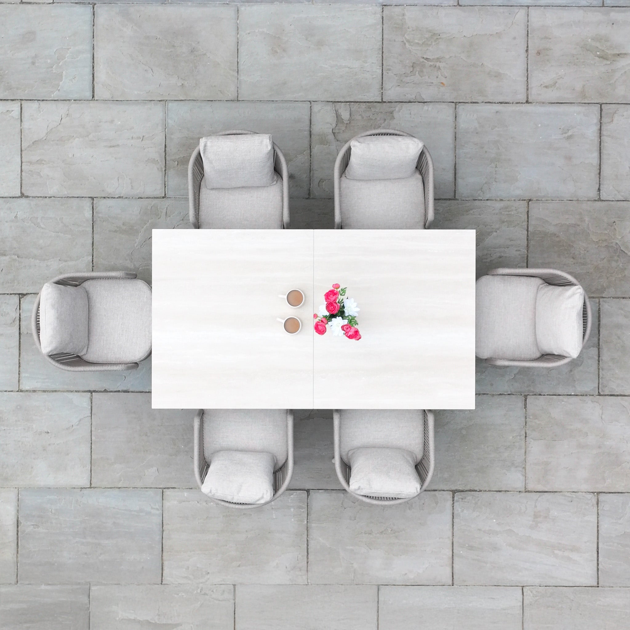 Top view of a Dekota 6 Seat Rope Dining Set with Albatross Extending Table in Fawn, set for two coffees and flowers, surrounded by six gray chairs on stylish stone tiles.