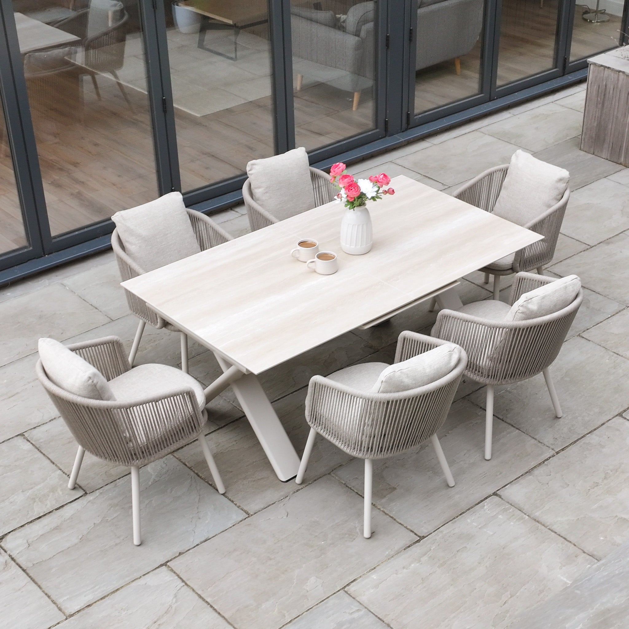 Enjoy outdoor meals with the Dekota 6 Seat Rope Dining Set featuring cushioned chairs and the Albatross Extending Table in Fawn, plus three mugs and a vase with pink flowers.
