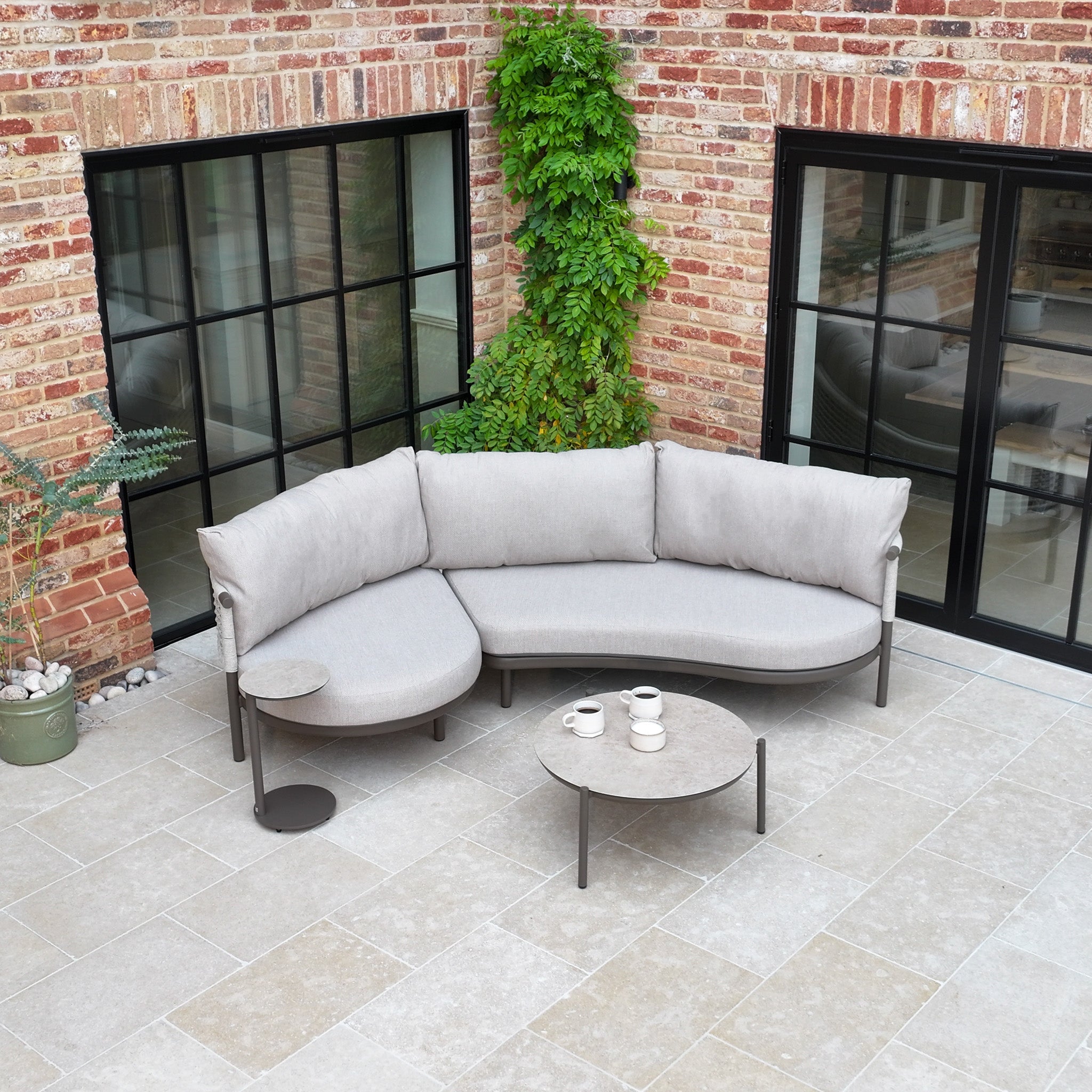 The Sutera Curved Chaise Sofa Set in Putty, featuring woven rope details and all-weather cushions, is placed on a tiled patio beside brick walls, large windows, and climbing plants.