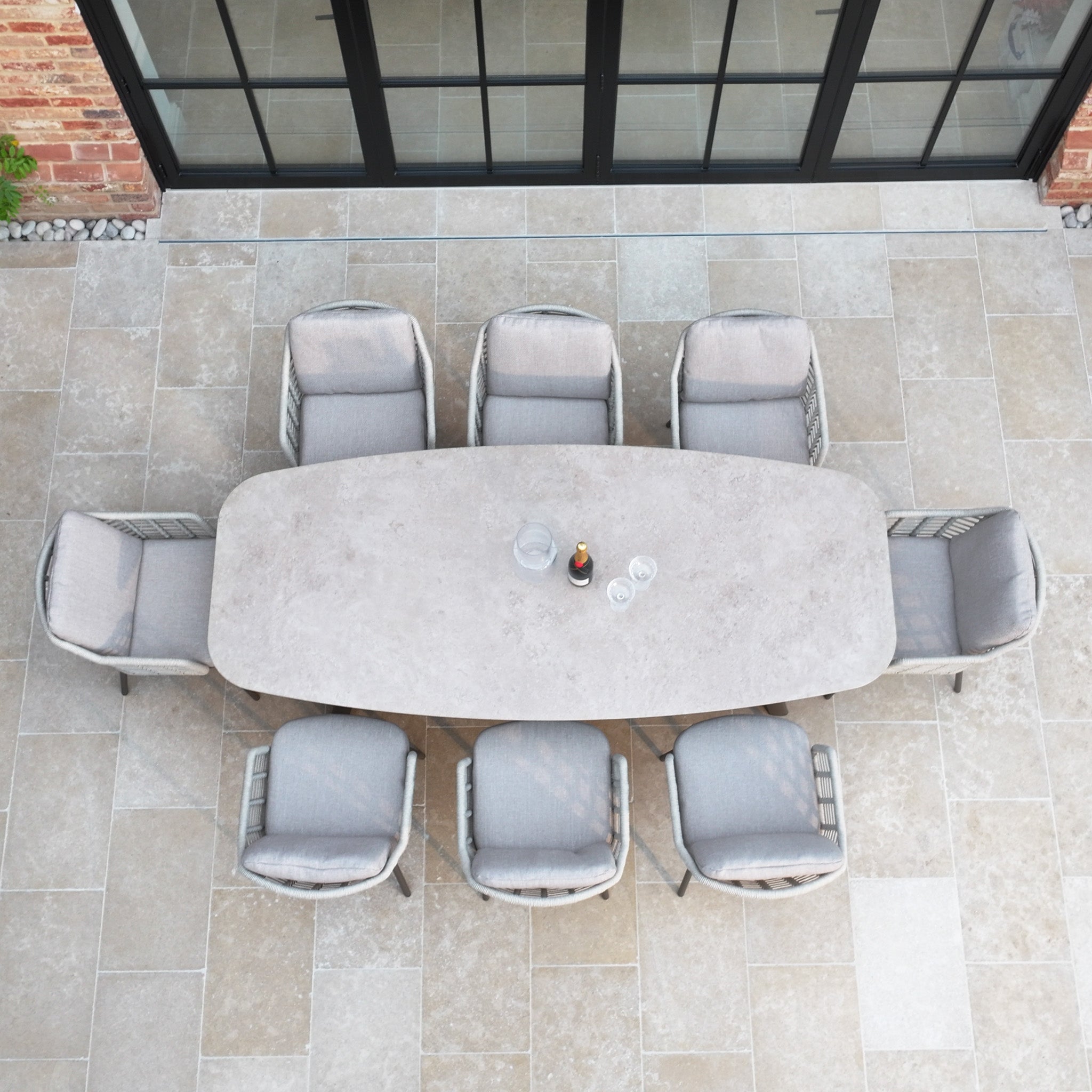 The Sutera 8 Seat Curved Rectangular Dining Set in Putty, viewed from above with eight gray chairs on a tiled patio near glass doors, offers a stylish setup perfect for outdoor gatherings.