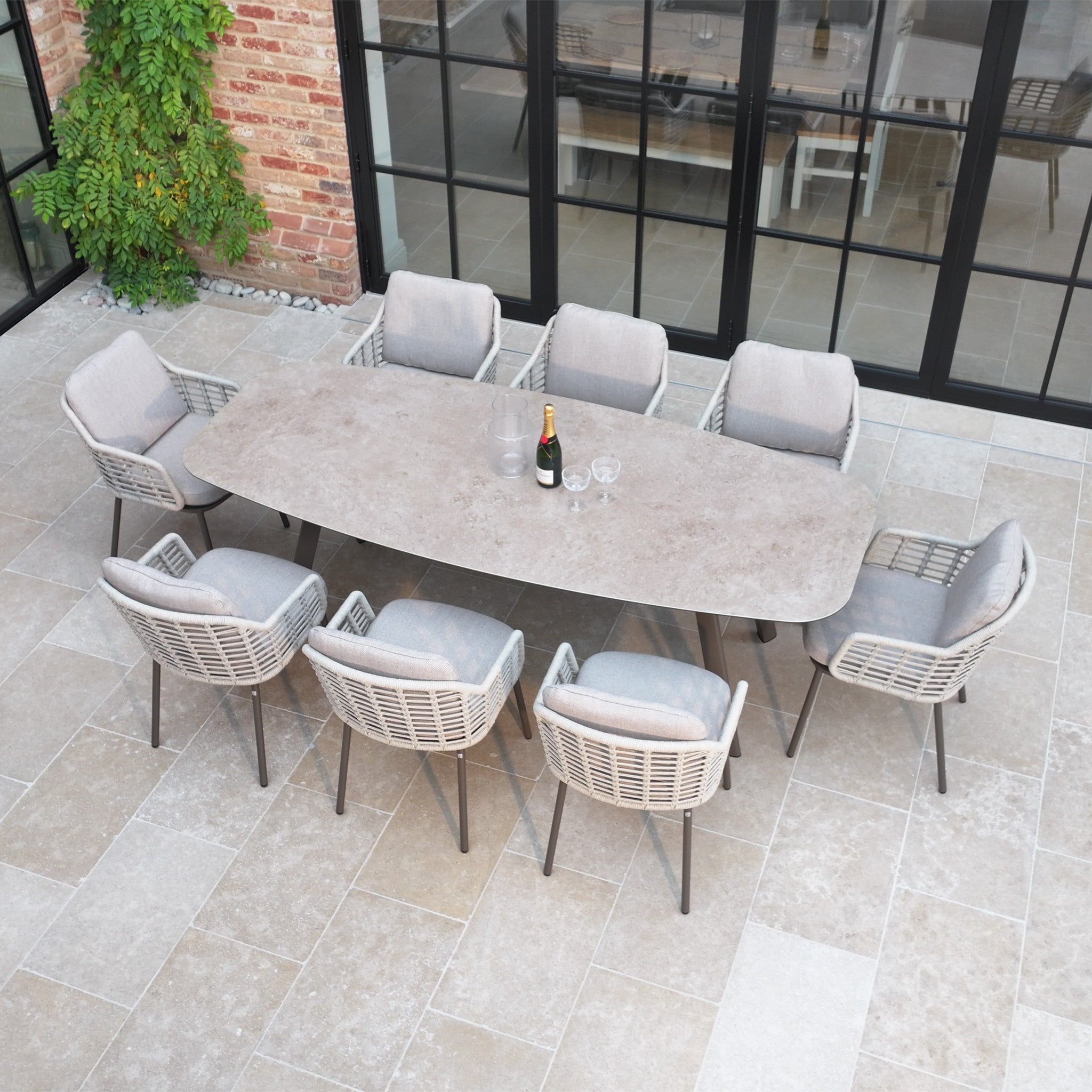 The Sutera 8 Seat Curved Rectangular Dining Set in Putty features cushioned chairs and is shown with a champagne bottle and glasses on a tiled patio, offering elegant outdoor dining as part of the Sutera garden furniture collection.