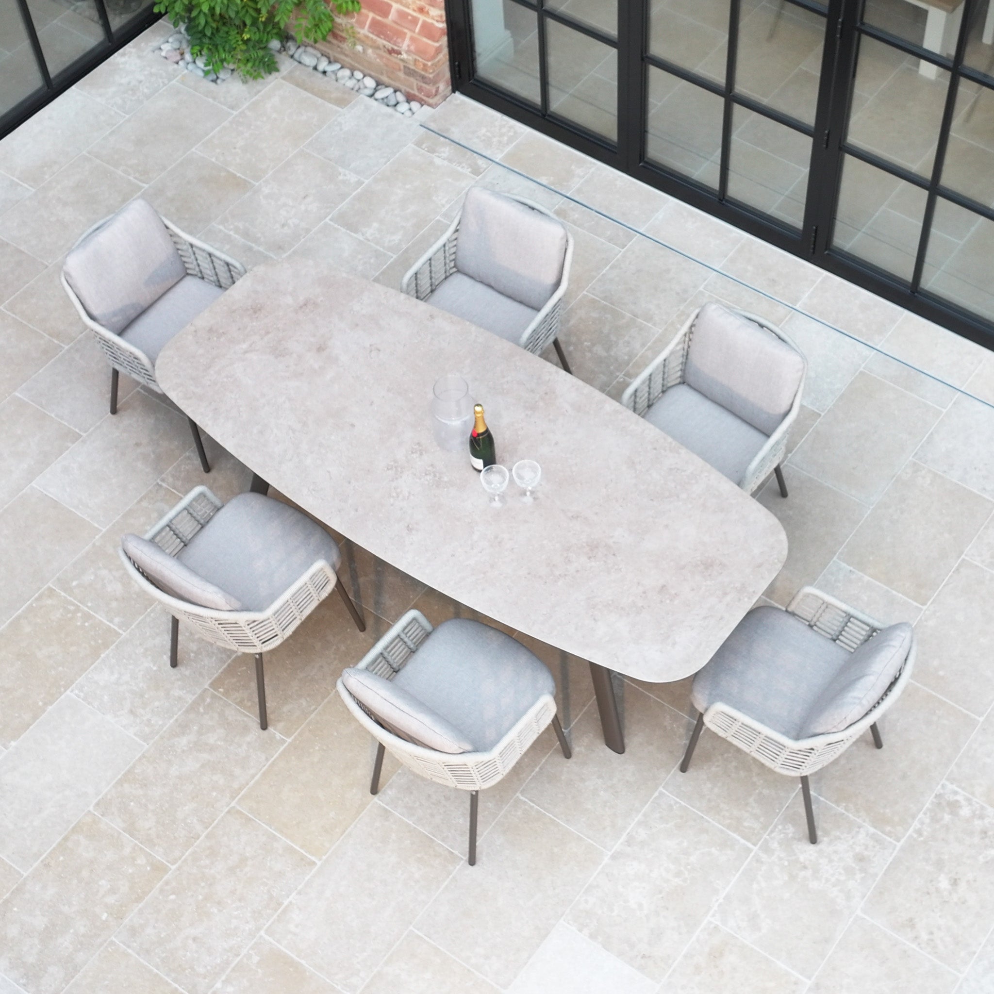 The Sutera 6 Seat Curved Rectangular Dining Set in Putty includes six cushioned chairs and an oval table, all crafted with all-weather fabric for year-round use. The table is styled with a wine bottle and glasses for elegant outdoor dining.