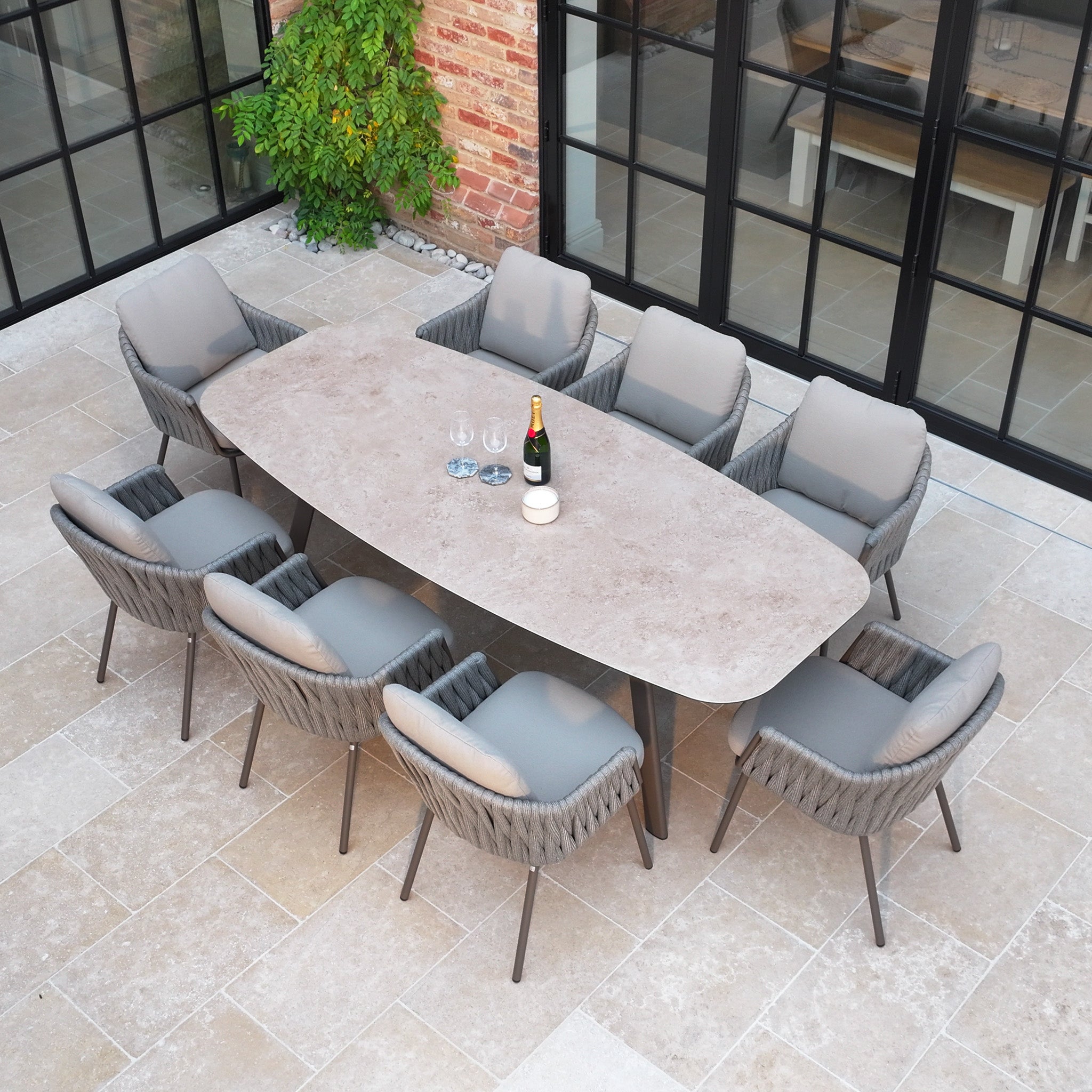 The Capri 8 Seat Curved Rectangular Dining Set in Stone Grey, featuring eight cushioned chairs and a stone table, is elegantly arranged on a tiled patio near glass doors—perfect for elevating outdoor gatherings.
