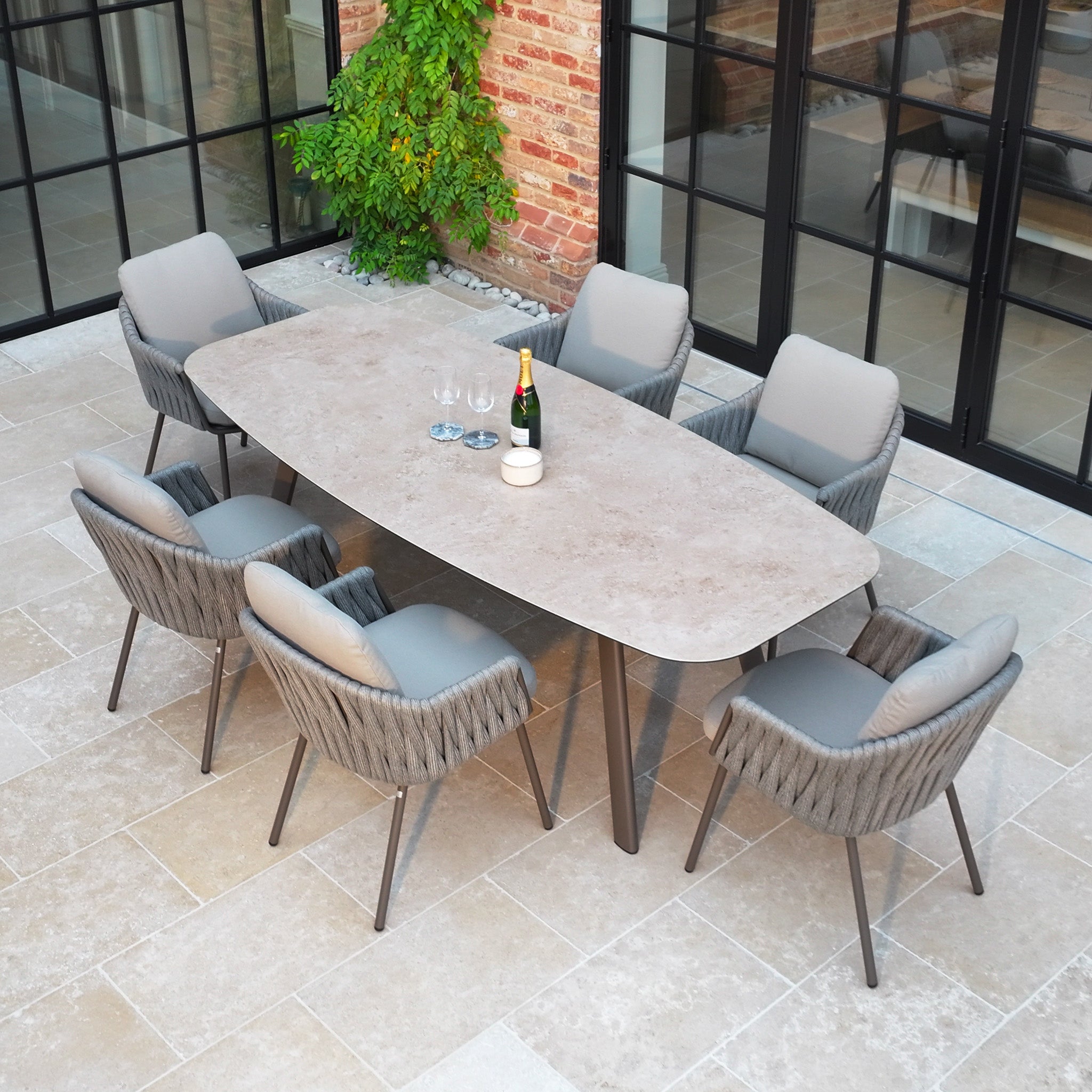 Enjoy outdoor dining with the Capri 6 Seat Curved Rectangular Dining Set in Stone Grey, featuring hand-woven rope detailing and six cushioned chairs, set beside a brick and glass building.