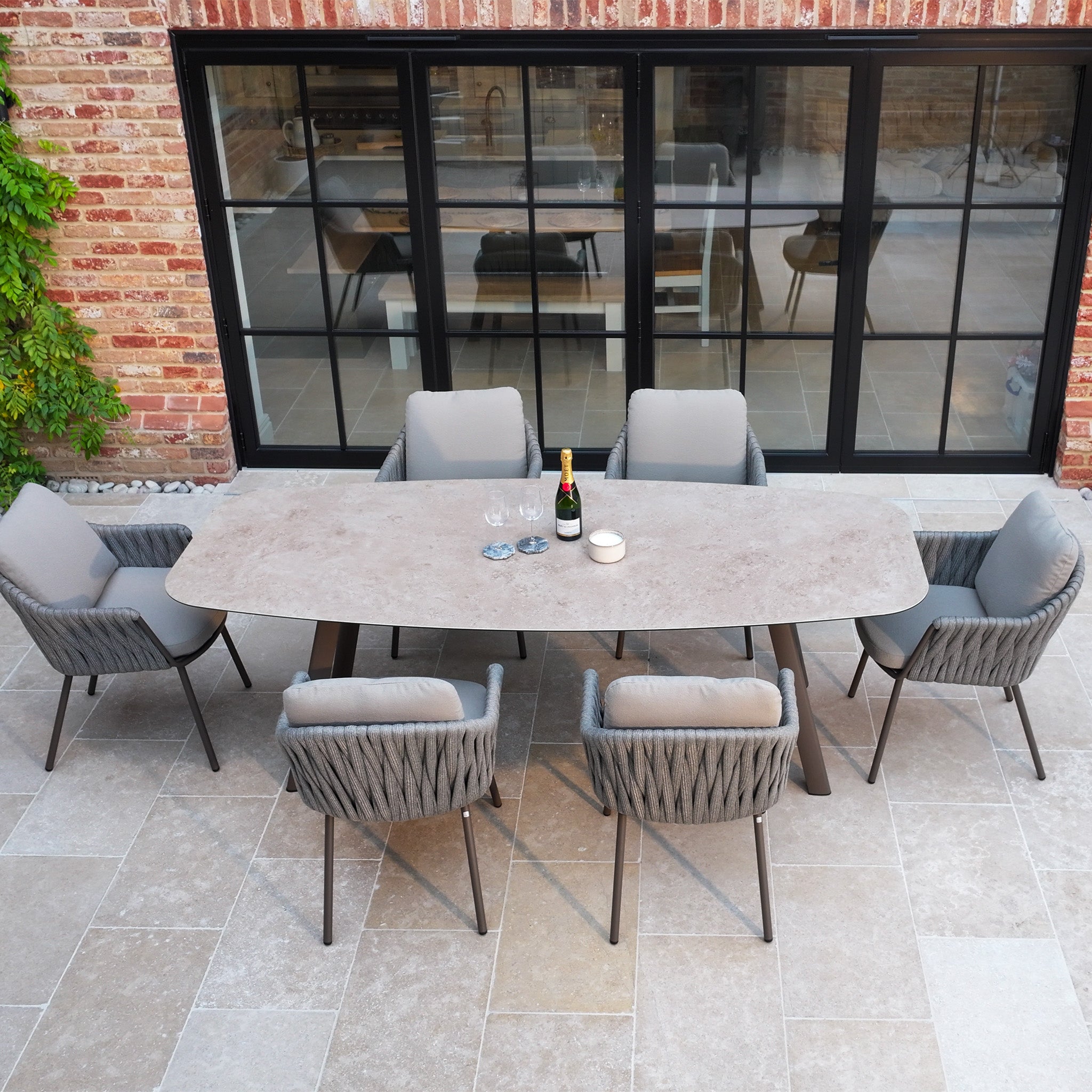 The Capri 6 Seat Curved Rectangular Dining Set in Stone Grey features six cushioned chairs and a table with hand-woven rope detailing, perfect for outdoor spaces like patios by glass doors.