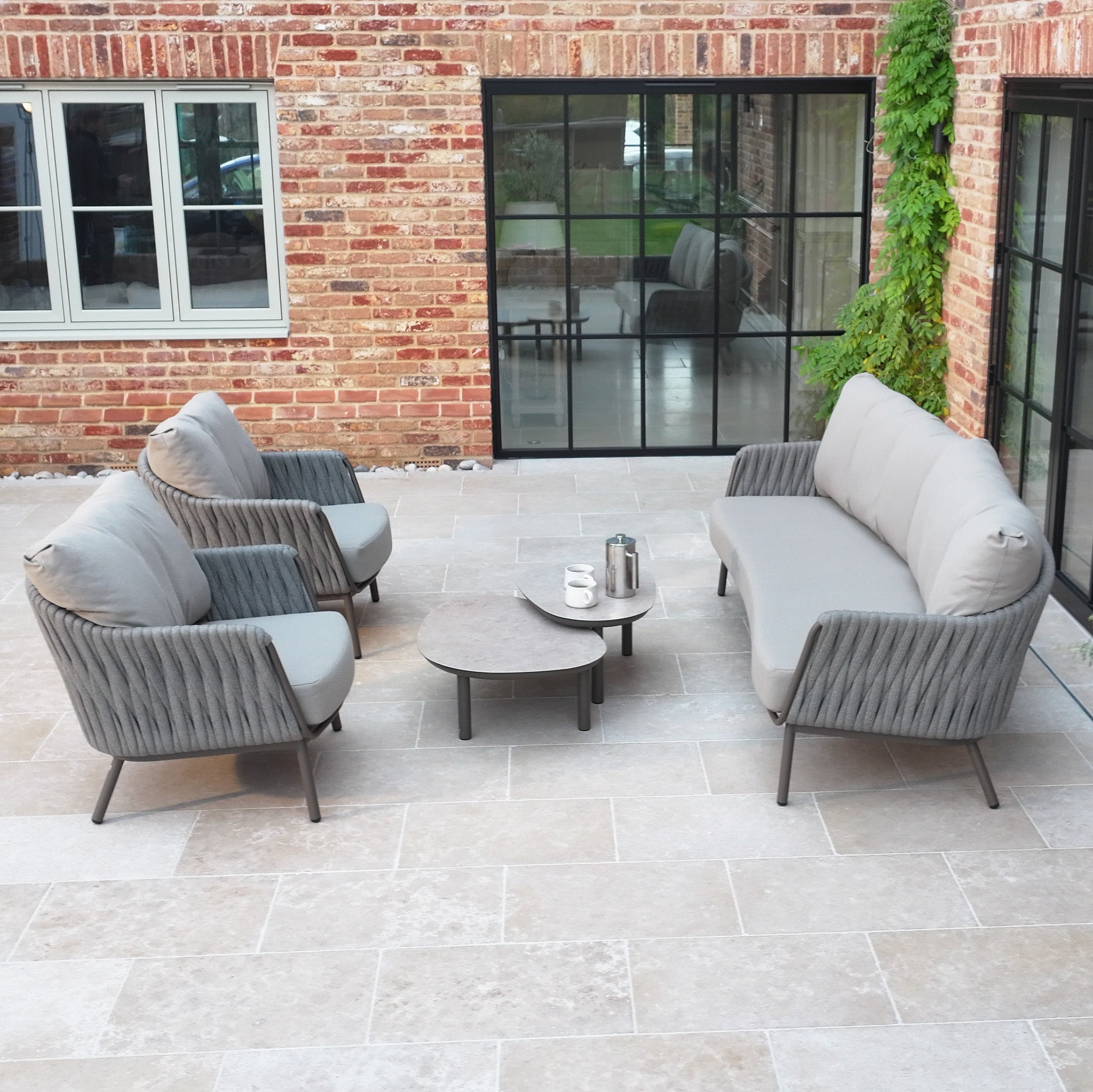 The Capri 3 Seat Sofa Set in Stone Grey features a sofa, gray cushioned chairs, and a coffee table with elegant hand-woven rope detailing—perfect for modern outdoor stone terraces.
